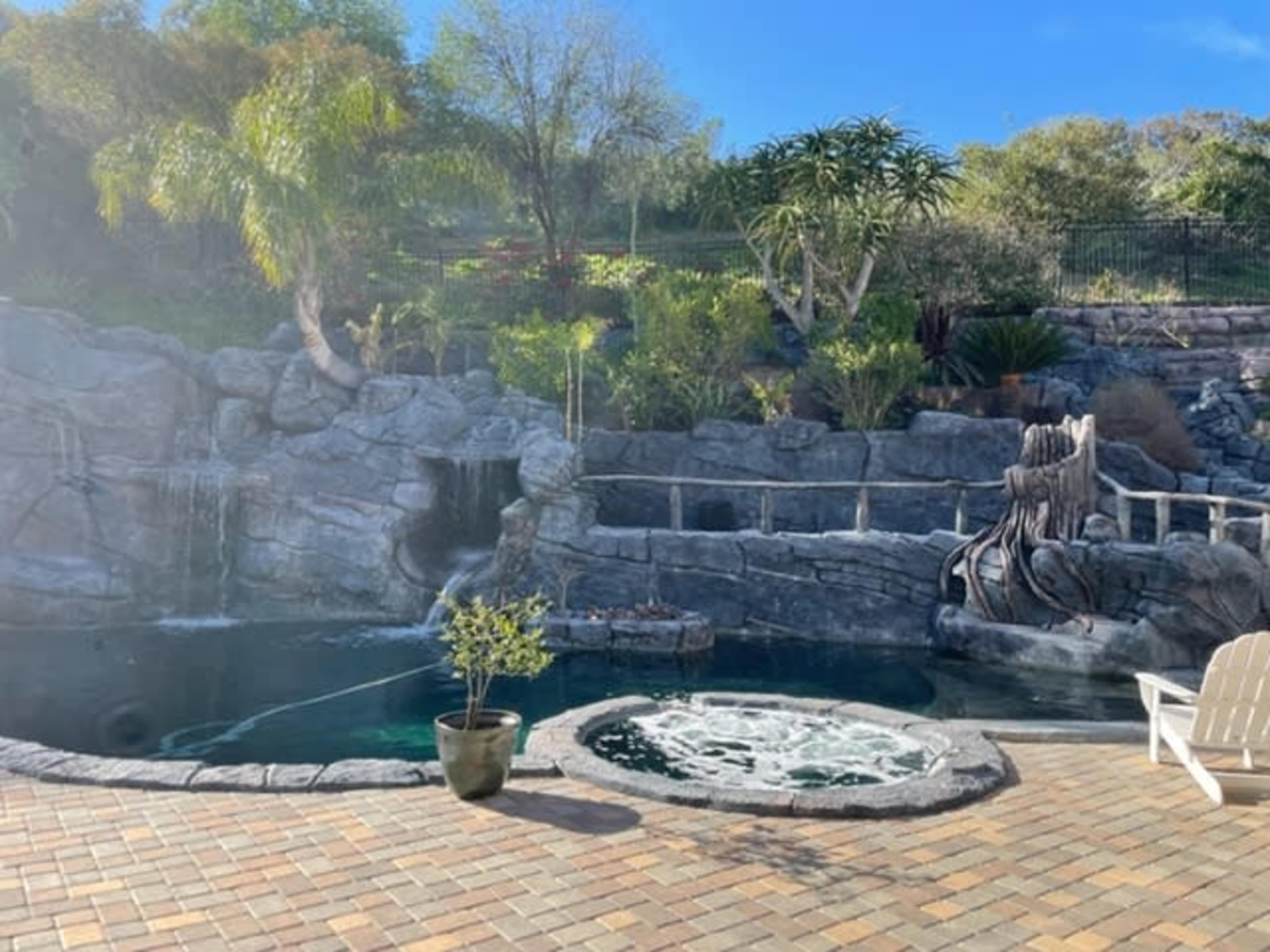 The image shows a landscaped backyard with a rock-themed swimming pool, a waterfall feature, and a hot tub surrounded by greenery.