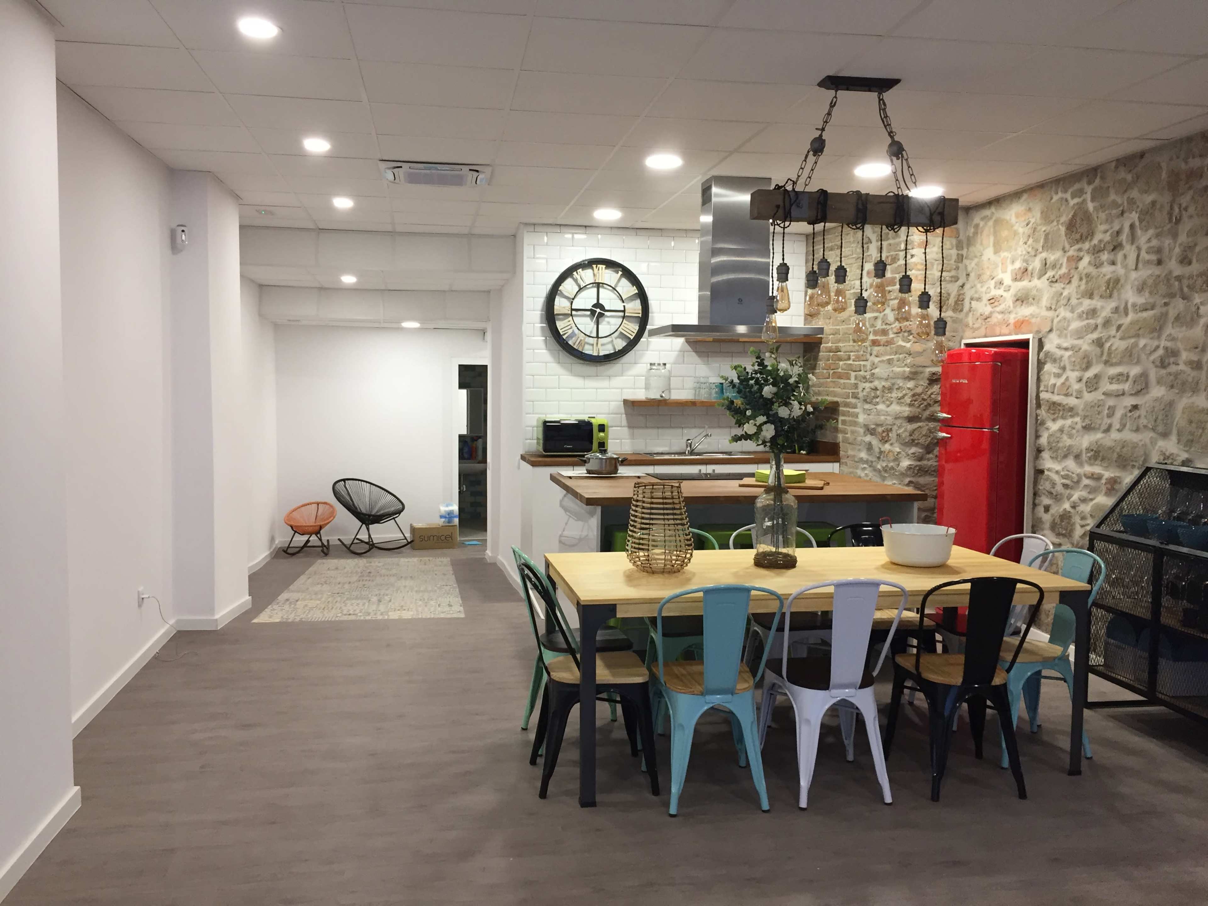 A modern kitchen and dining area features a large table with multicolored chairs, a red refrigerator, and a stone wall backdrop.