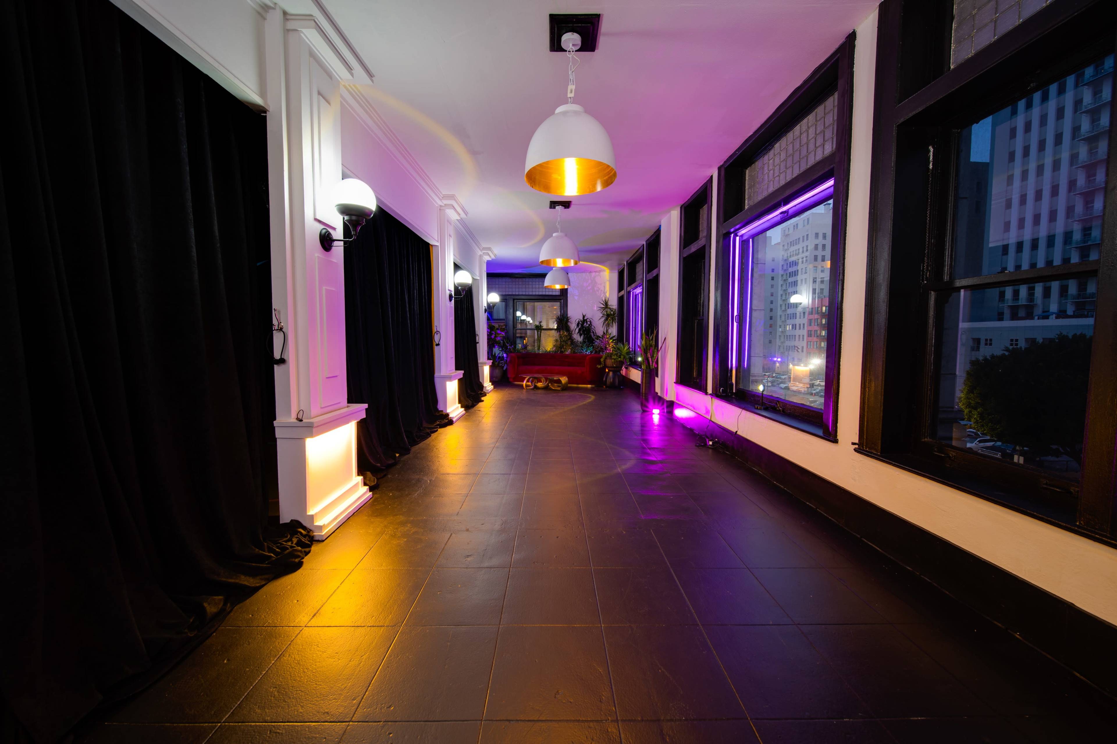 A well-lit corridor featuring black curtains, stylish light fixtures, and large windows, with ambient lighting creating a modern atmosphere.