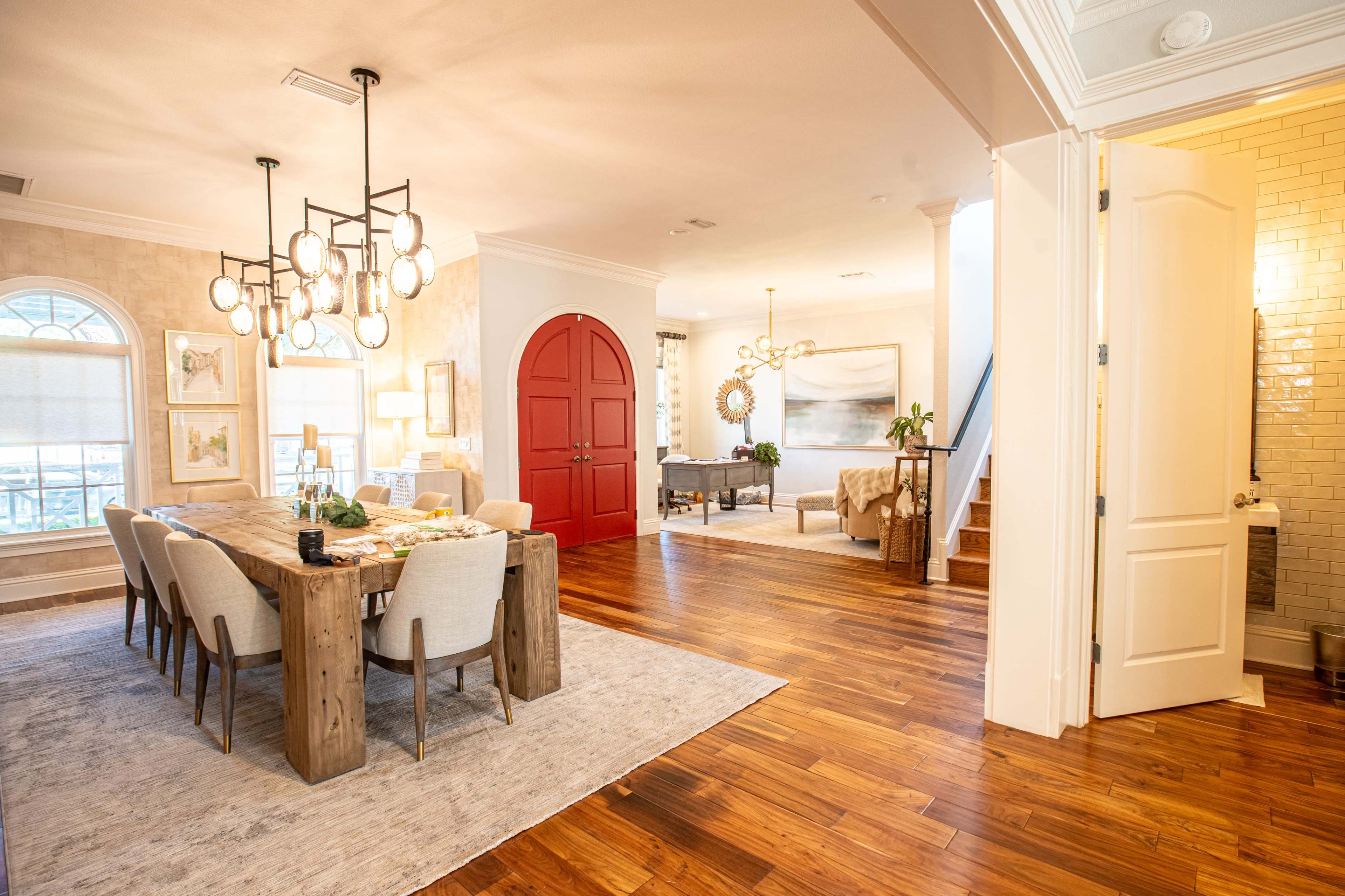 The image shows a well-lit dining area with a wooden table surrounded by chairs, open to a living room and a staircase, featuring a bright red door on the right.
