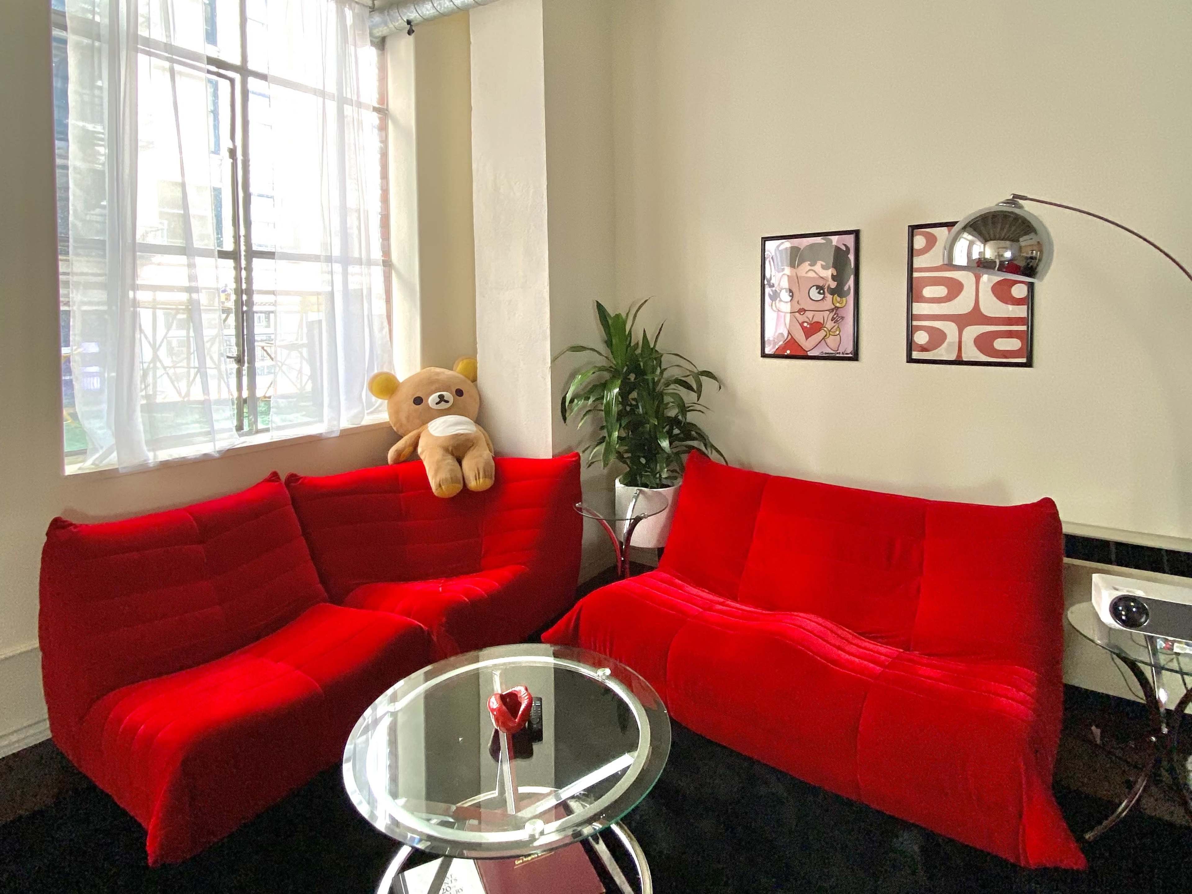 A bright room features red sofas, a glass coffee table with a decorative item, a plush toy sitting on one sofa, and framed artwork on the walls.