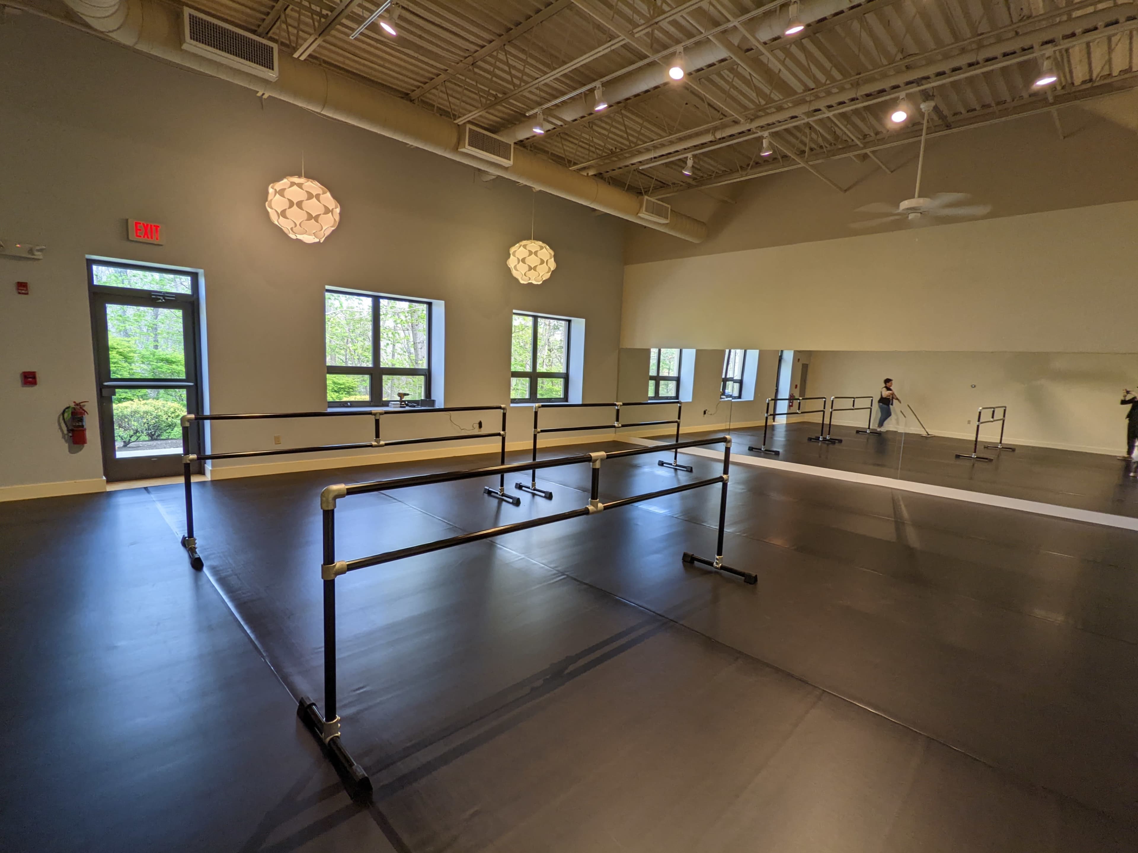 The image shows a bright dance studio with black flooring, mirrored walls, and ballet barres arranged along one side.