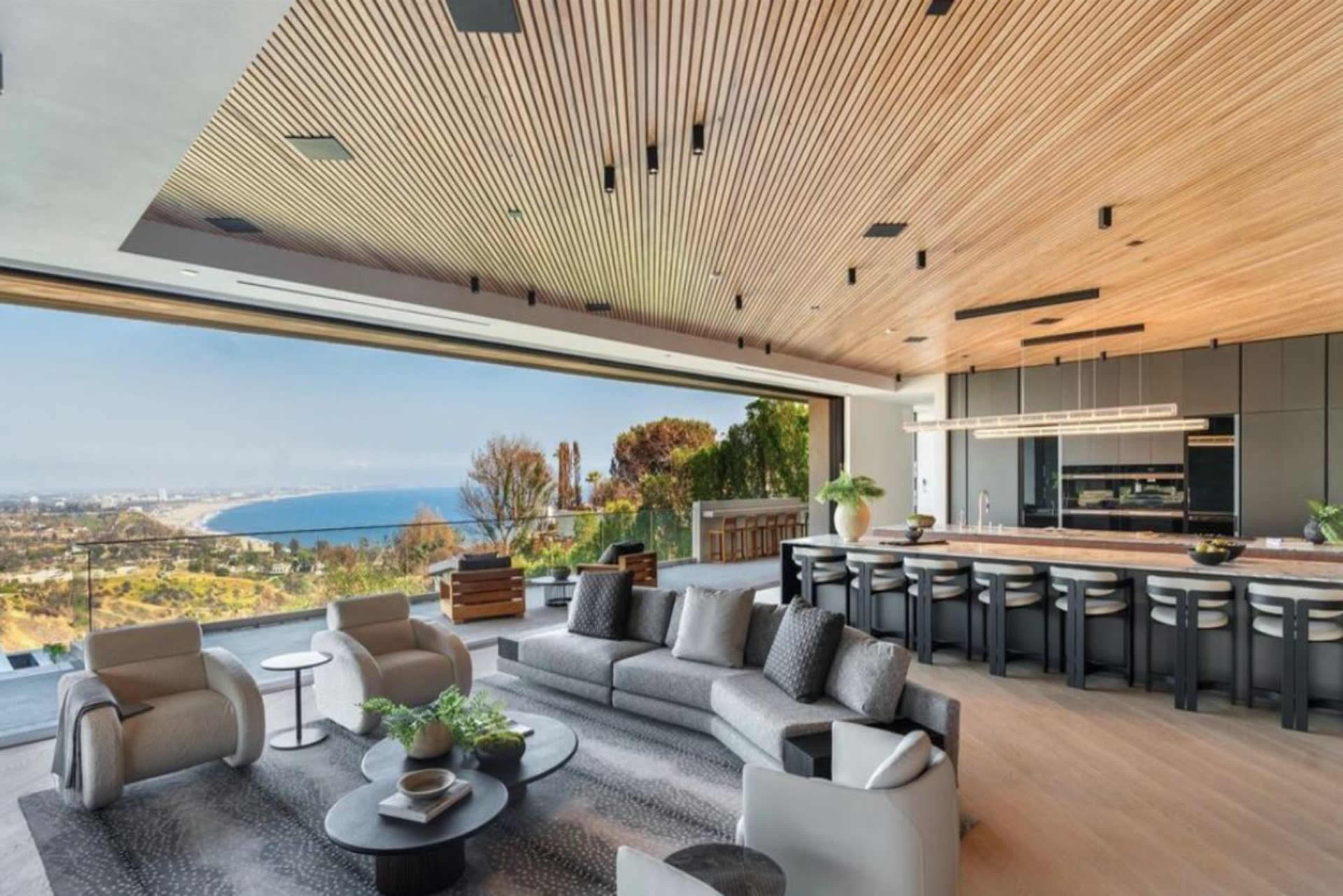 The image shows a modern open-concept living space featuring a large window with a view of the ocean, a seating area with a sofa and chairs, and a dining area with a kitchen in the background.