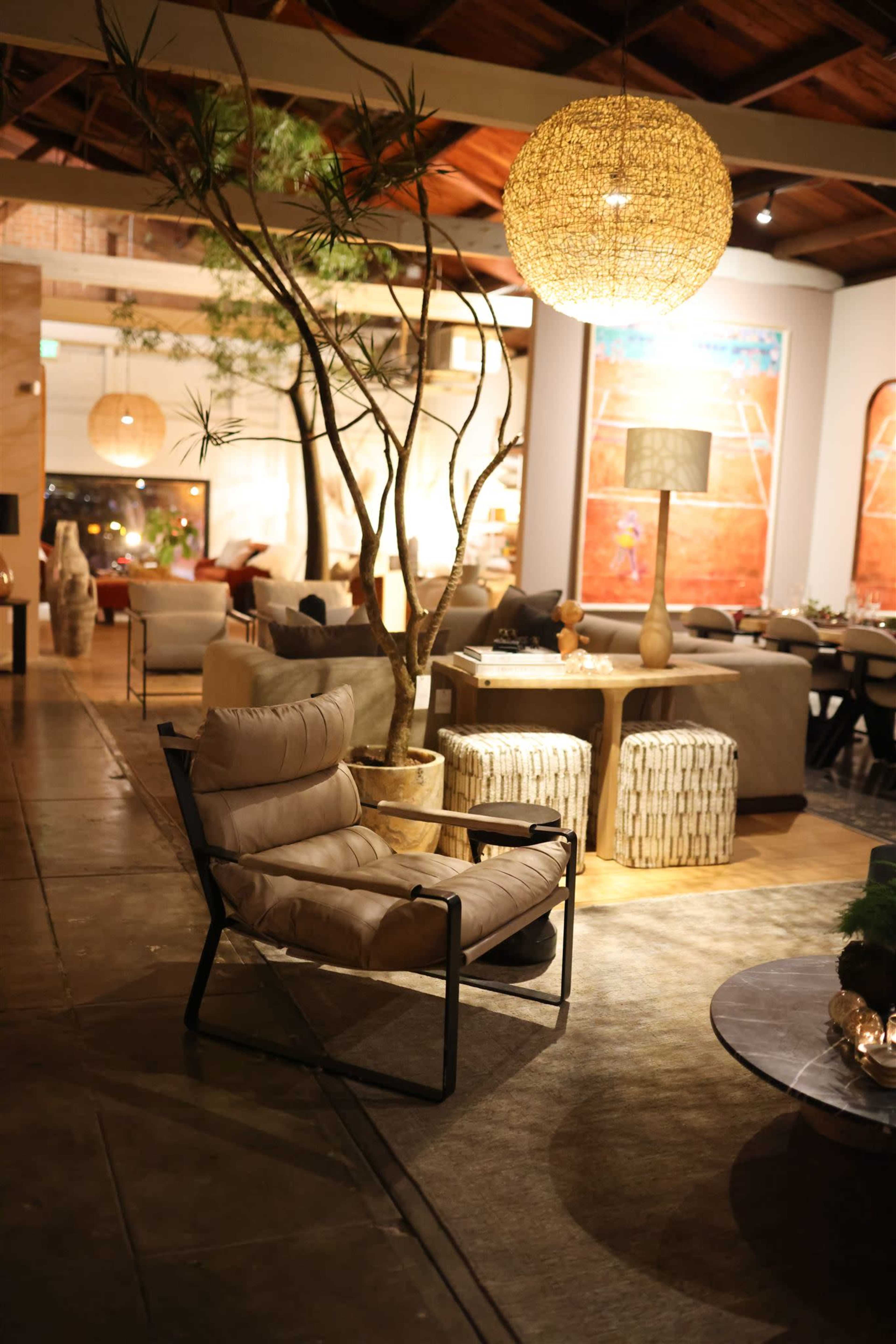The image shows a stylish, contemporary interior of a café or lounge featuring modern furniture, a large pendant light, and a decorative tree.