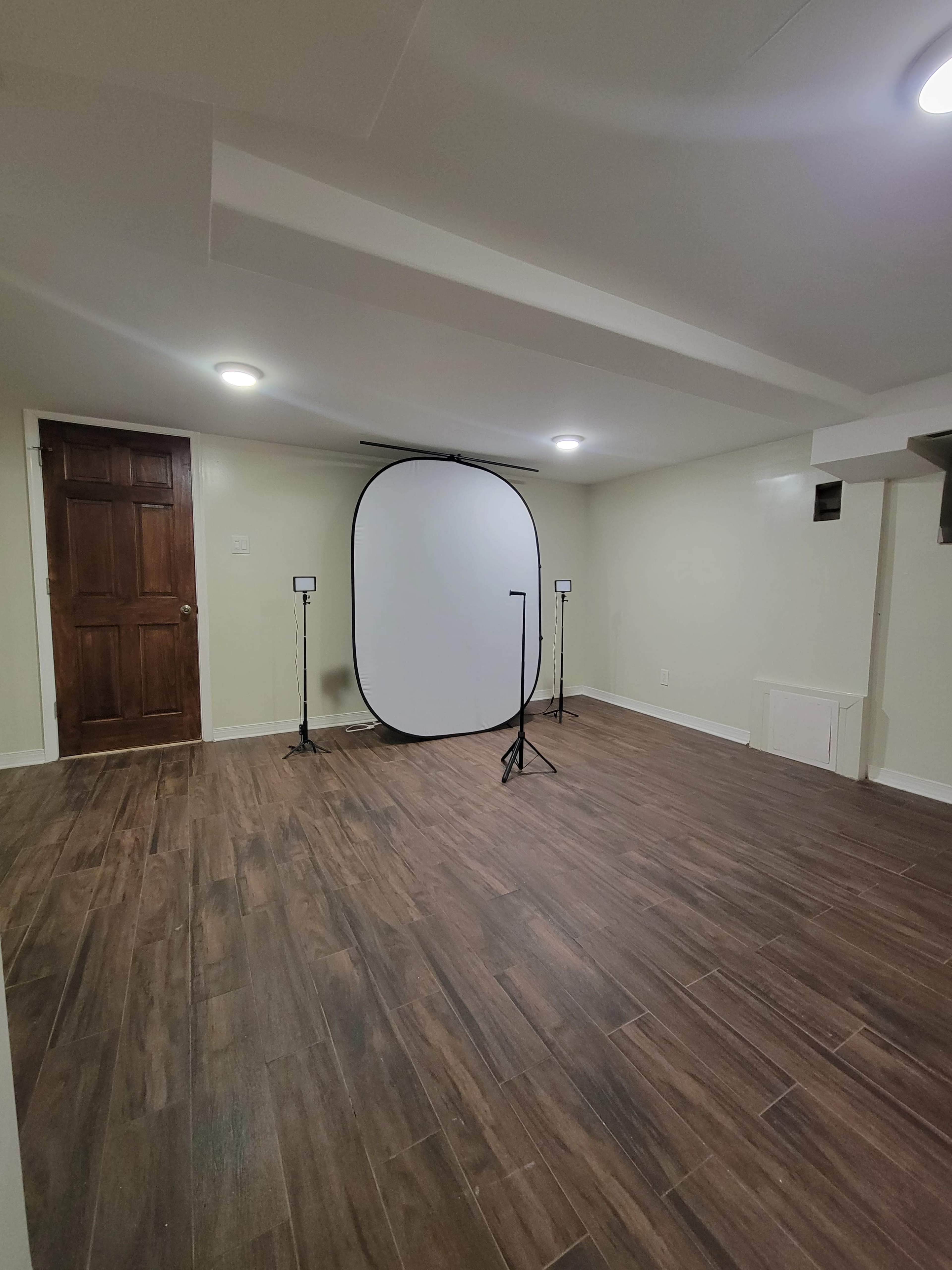 A spacious basement room features a large white backdrop on a stand, two lighting fixtures, and a wooden door.