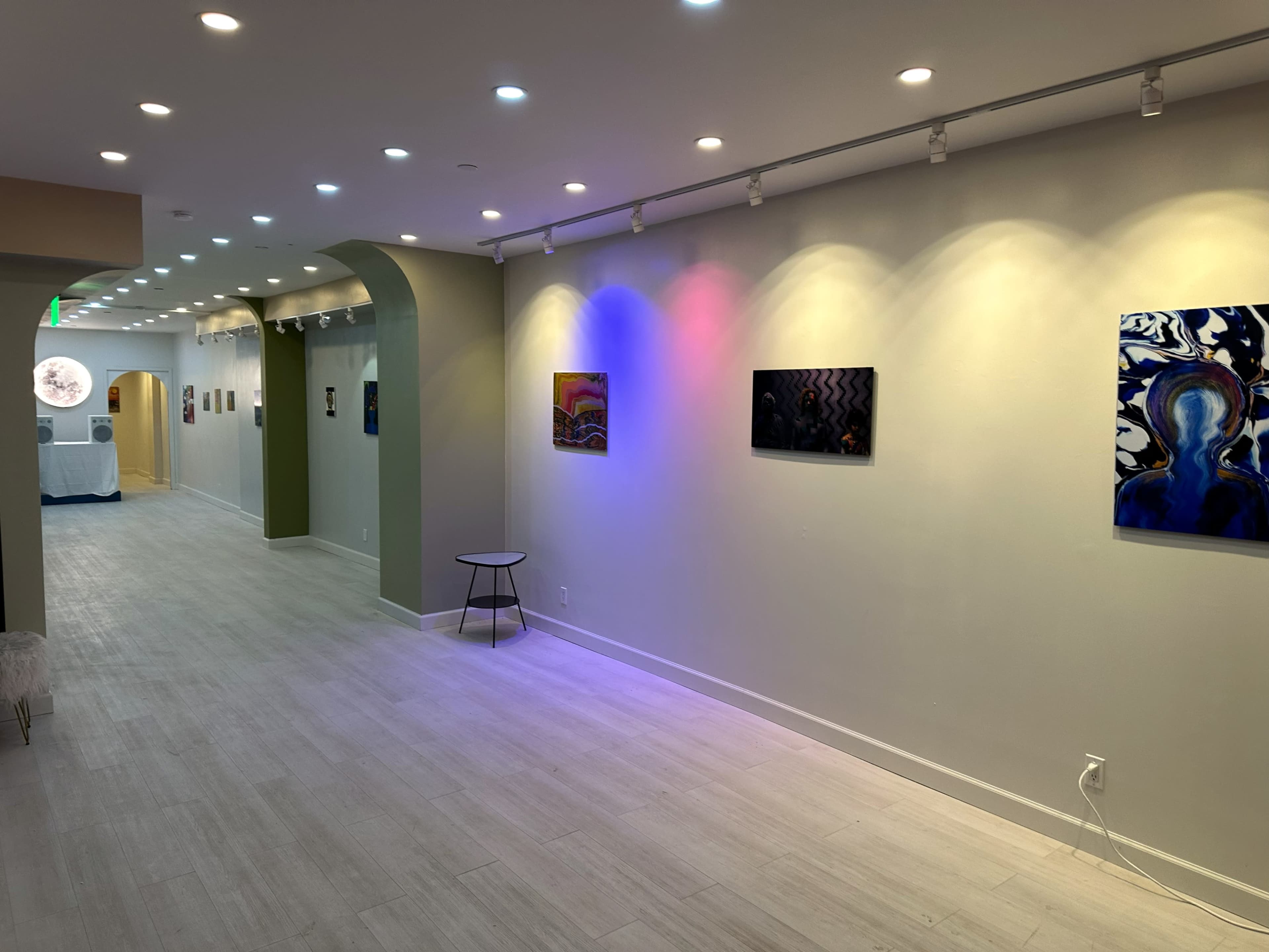 The image shows an art gallery with a long hallway featuring several colorful paintings illuminated by track lighting on the walls.
