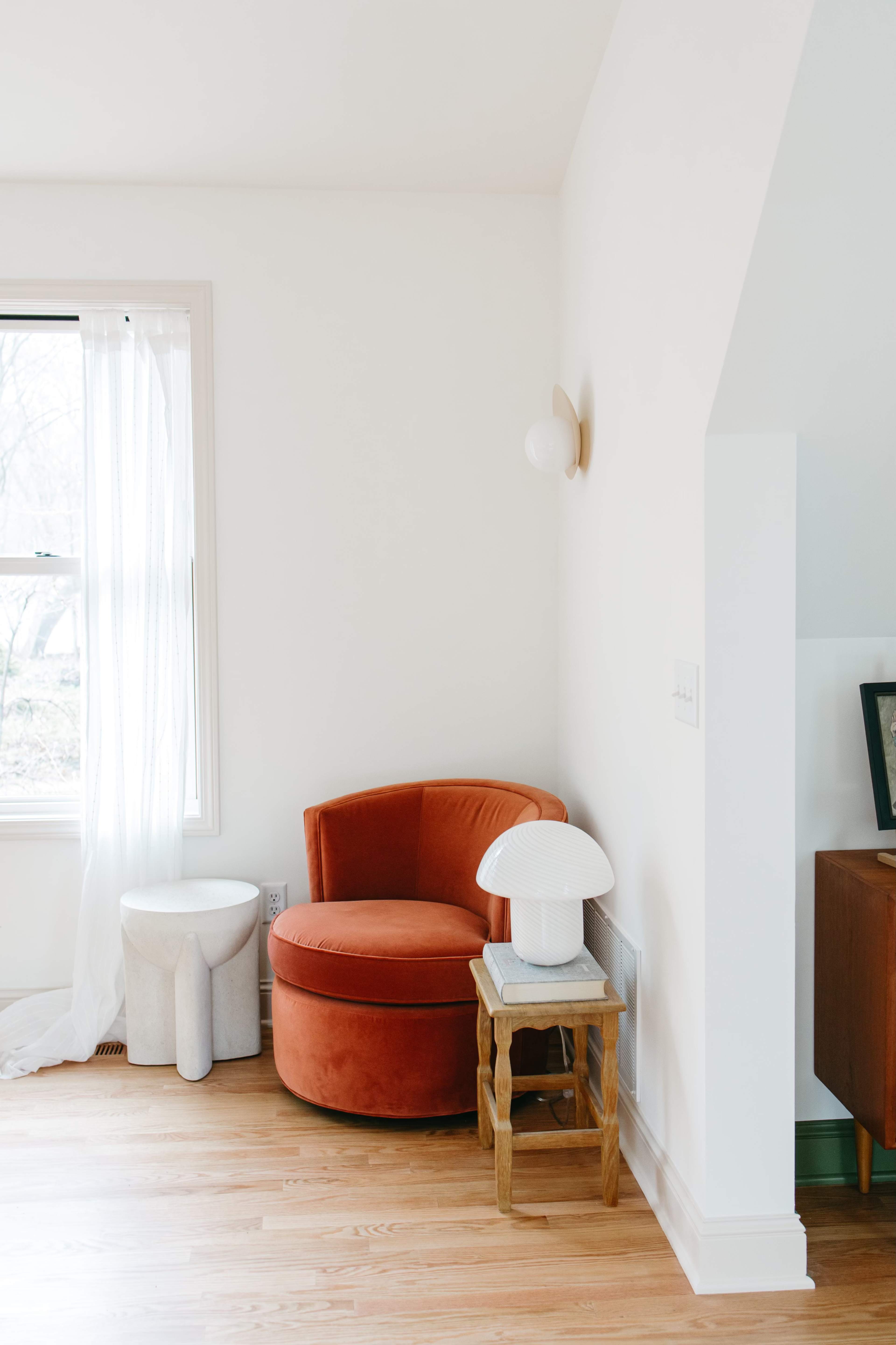 A round, orange upholstered chair is positioned near a window, accompanied by a small white lamp and a wooden side table in a light-filled room.