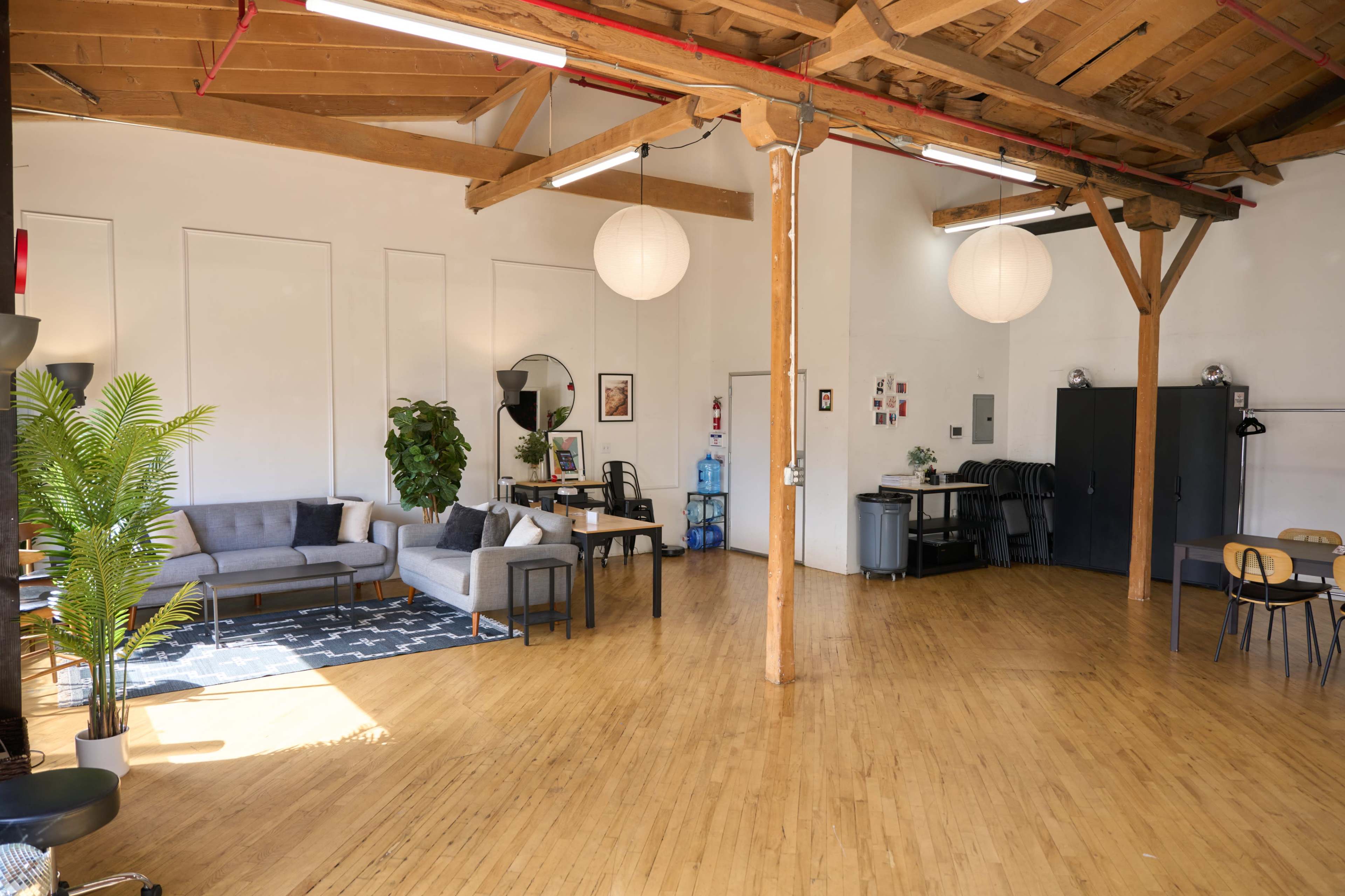 The image shows a spacious, well-lit interior of a modern loft with wooden beams, featuring a seating area with a couch, a dining space with chairs, and a plant in one corner.
