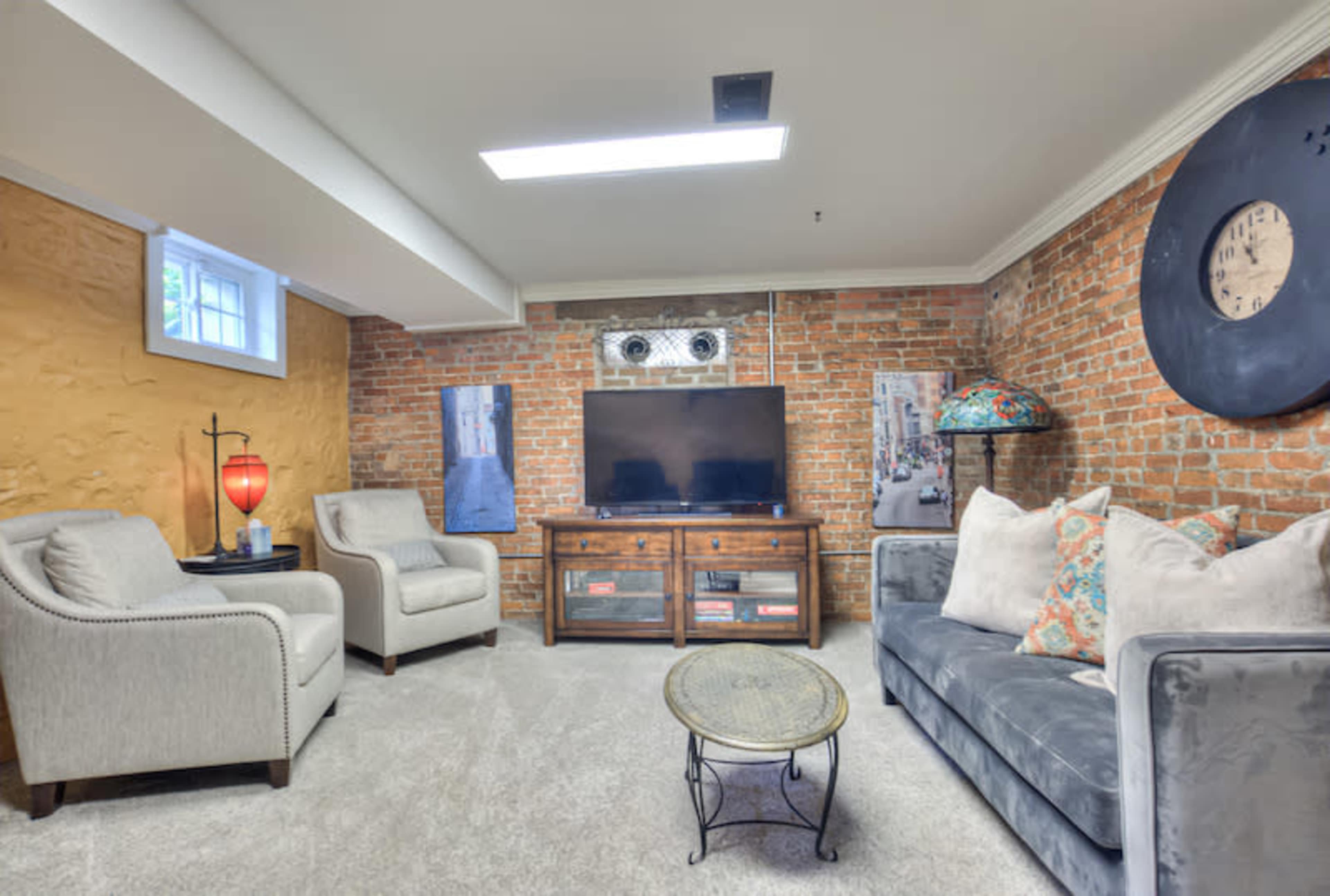 A basement living area with brick walls, a television on a wooden cabinet, two armchairs, and a sofa with decorative pillows.