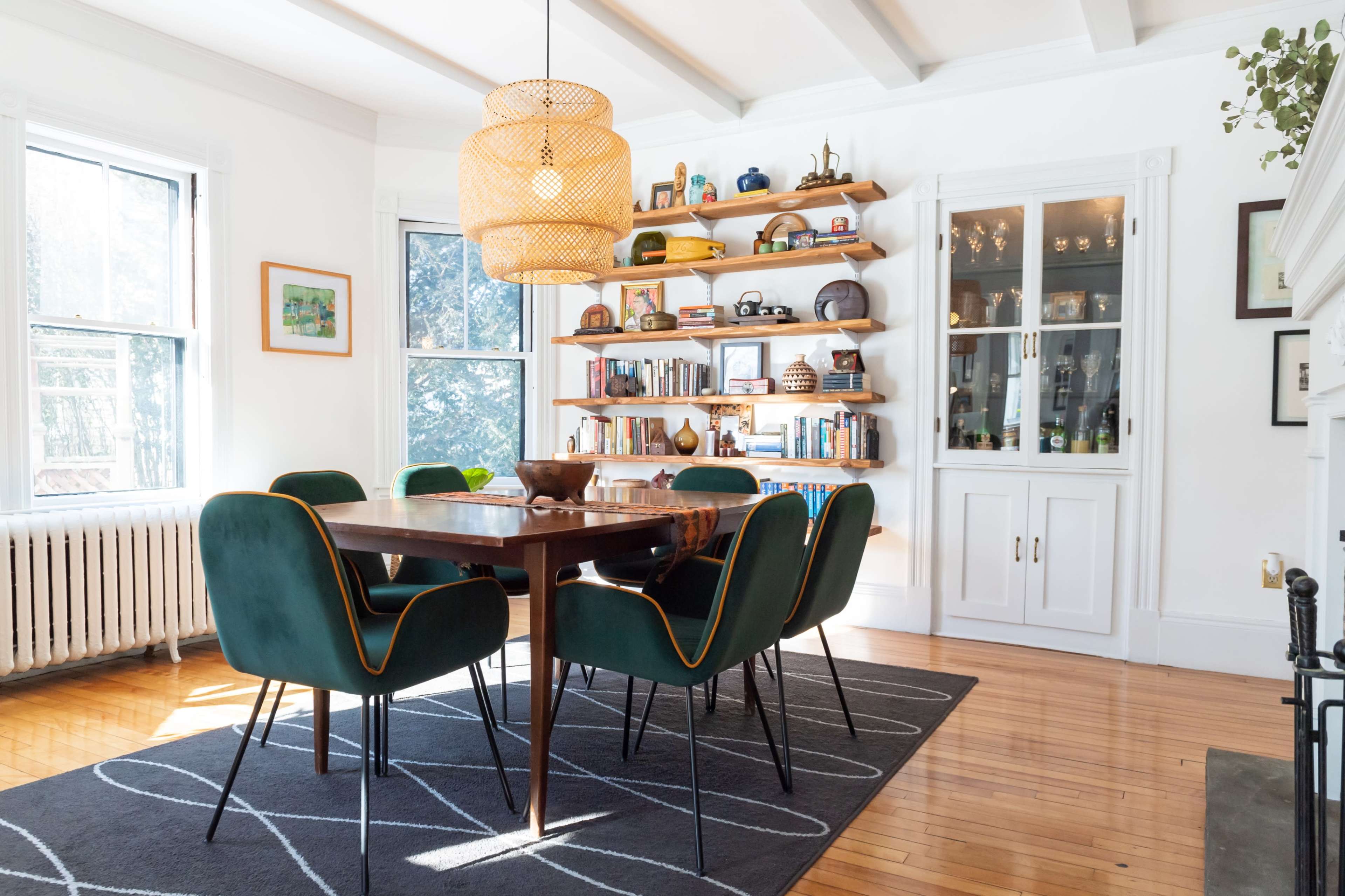A well-lit dining room features a wooden table surrounded by green chairs, with bookshelves displaying various items along the walls.