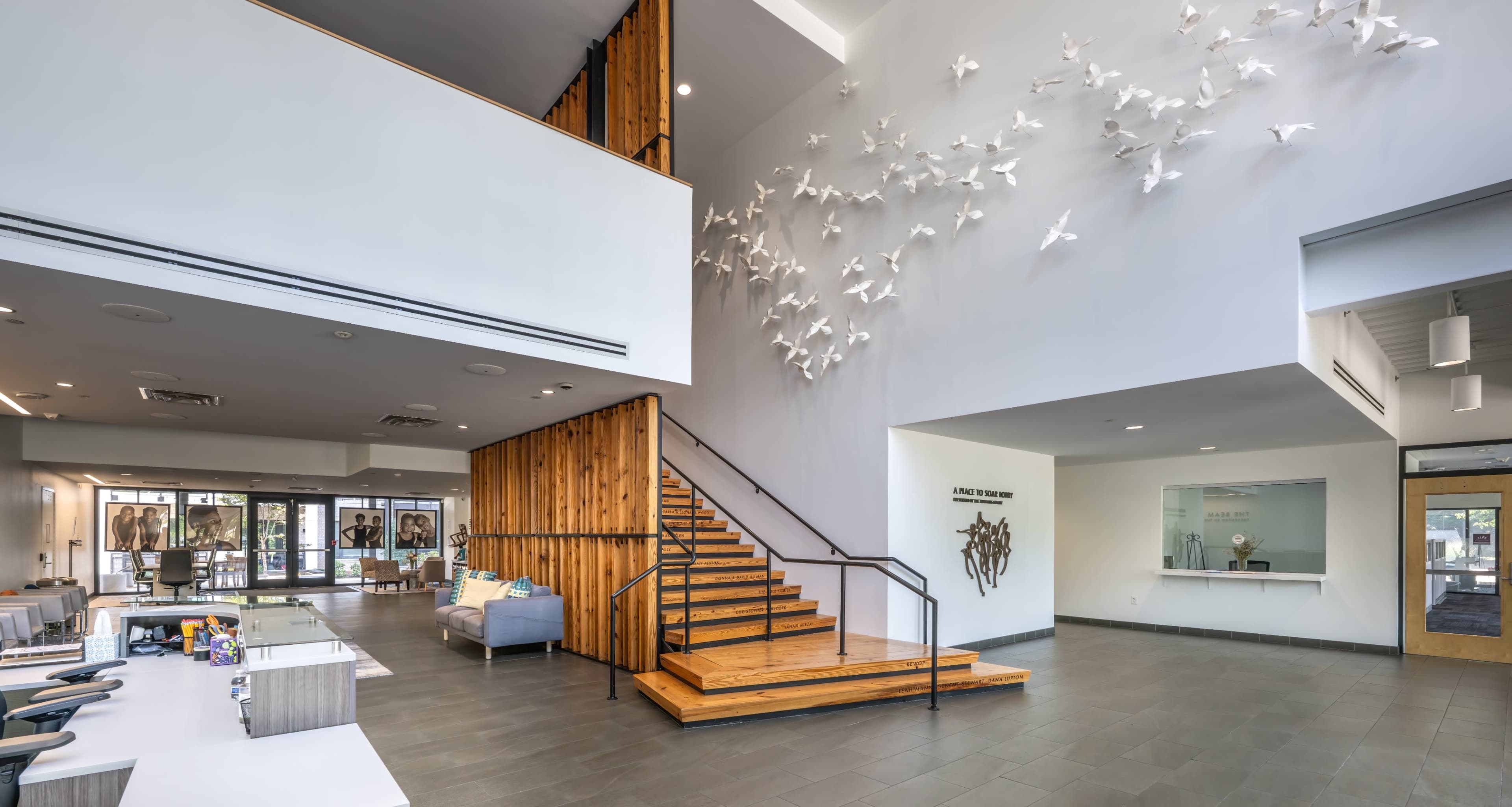 The image shows a modern reception area with wooden accents, a staircase, and a wall adorned with white decorative birds.