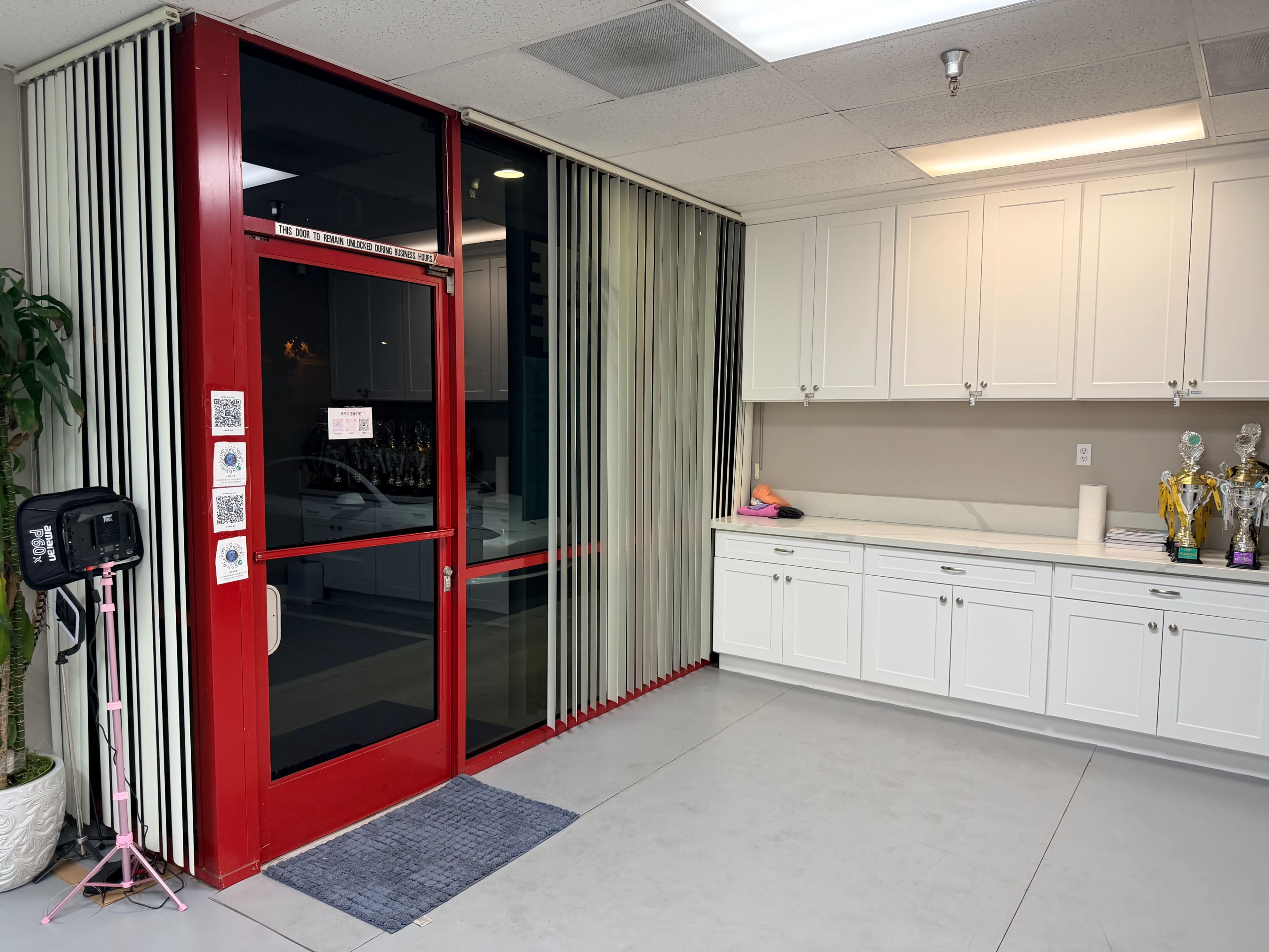 The image shows a commercial space with a red front door, vertical blinds, and white cabinetry along the wall.