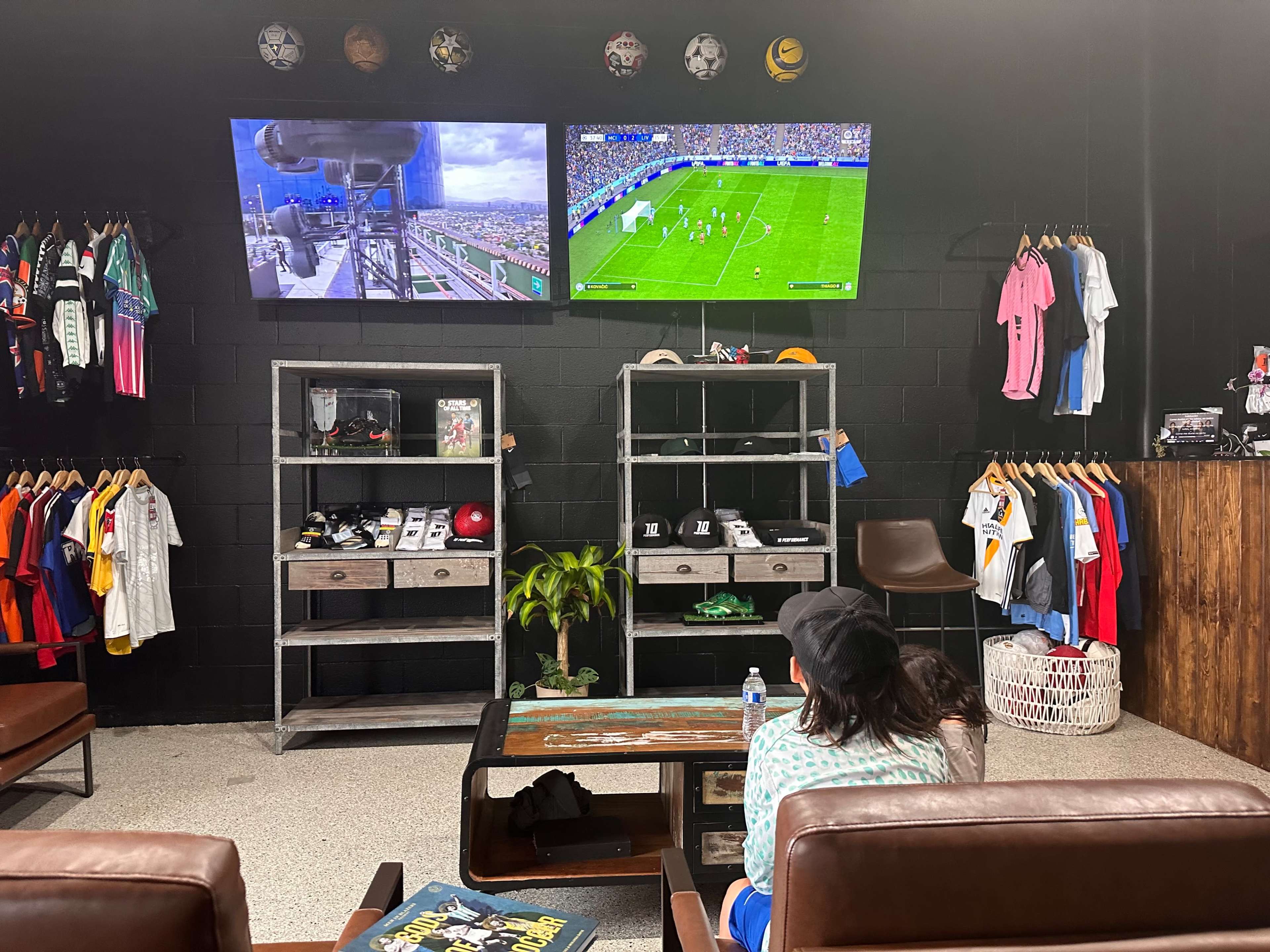 A sports-themed shop with two televisions displaying a soccer match, surrounded by racks of jerseys and sports merchandise.