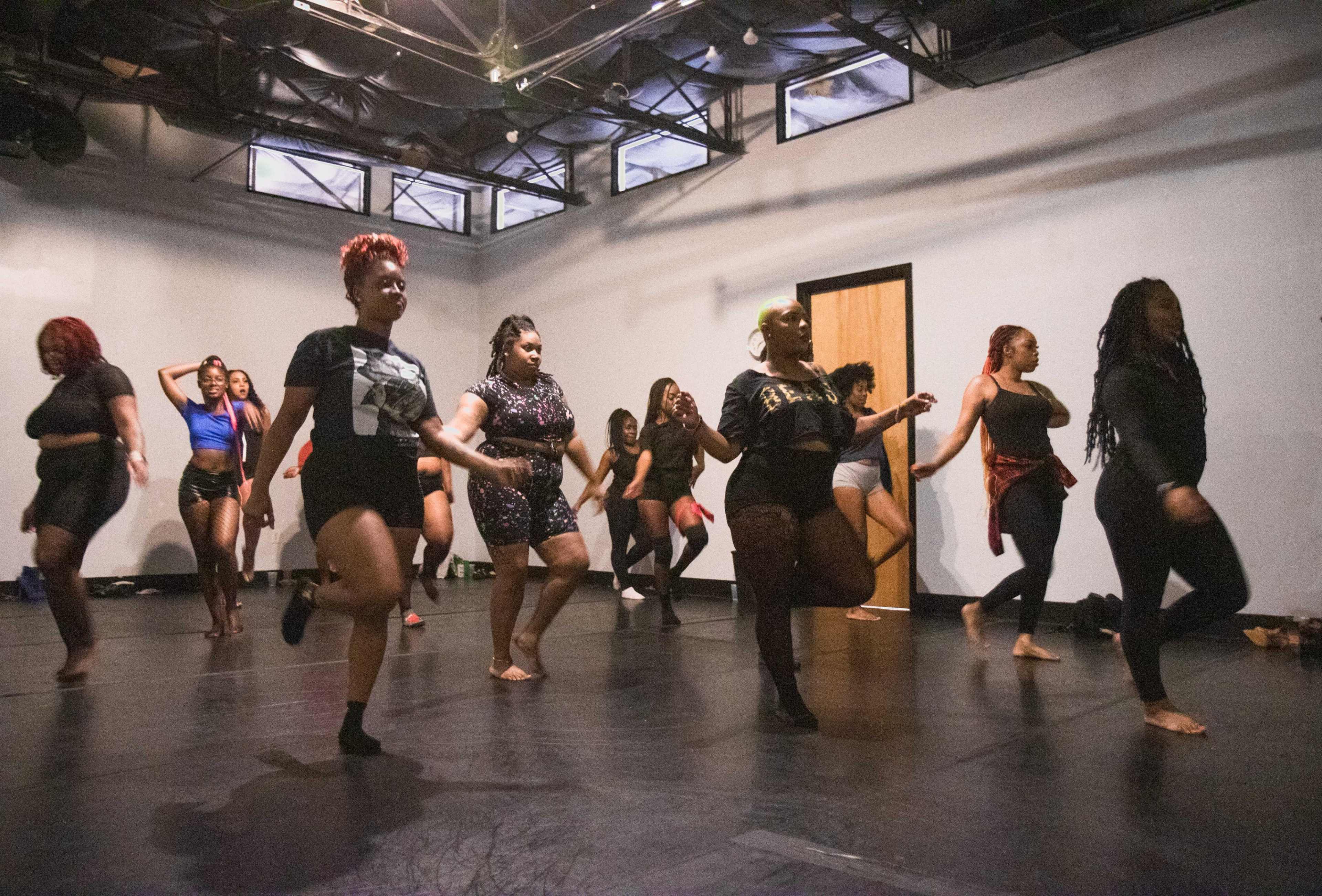 A group of women is practicing a dance routine in a studio with mirrored walls and wooden floors.