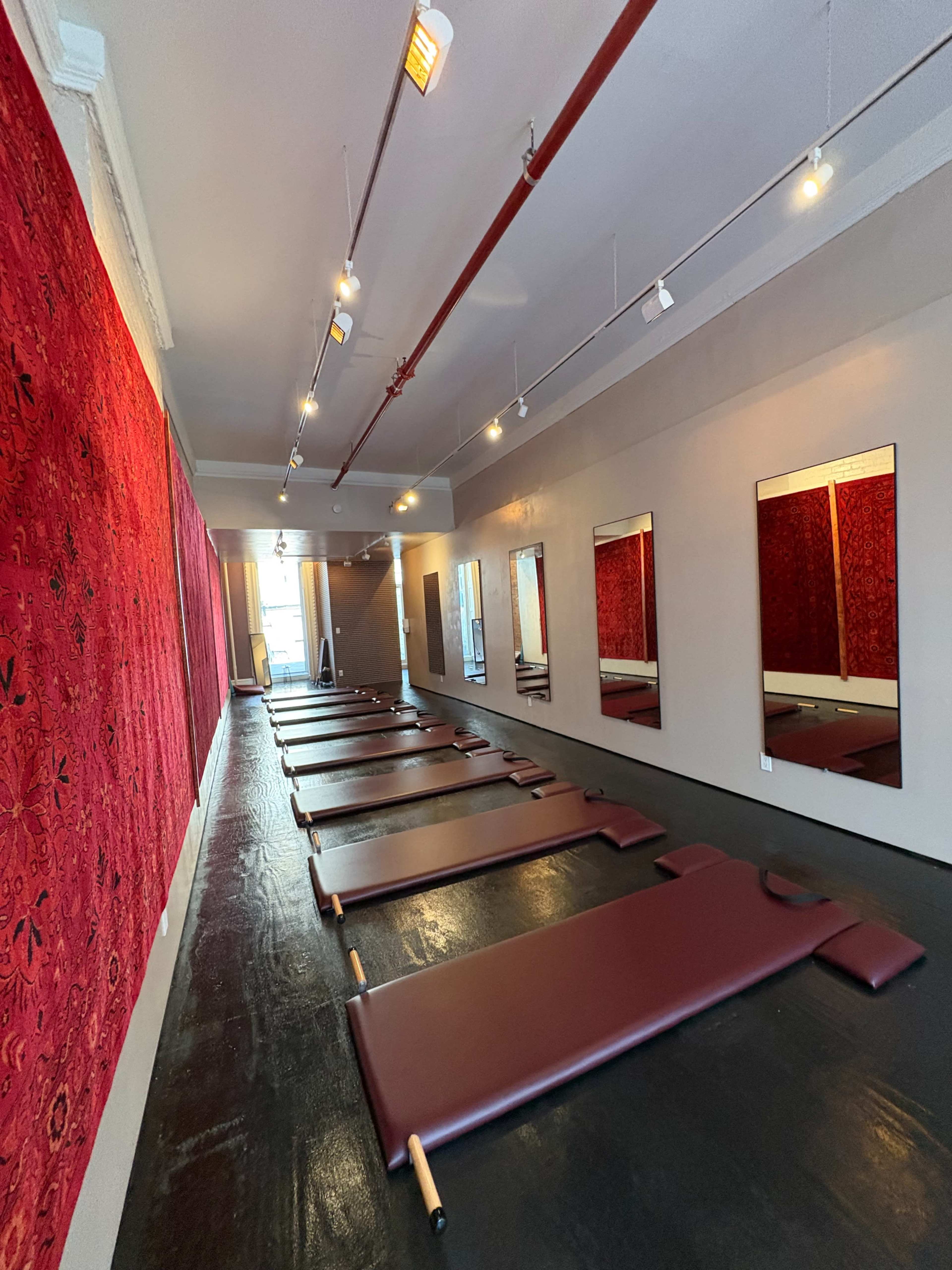 The image shows a sparsely furnished studio with maroon mats arranged on a dark floor, flanked by large mirrors and red patterned walls.