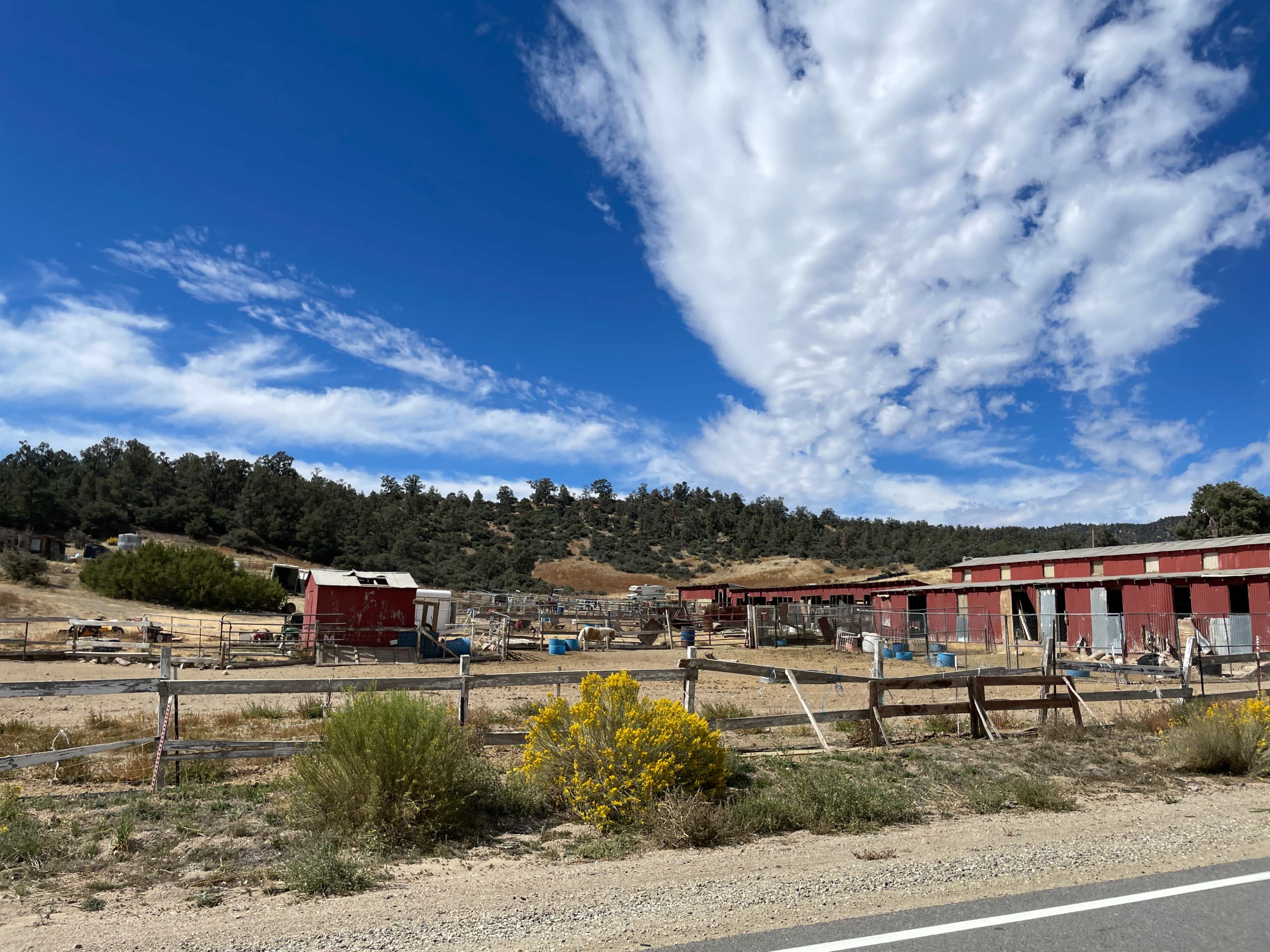 A rural scene features a barn and several outbuildings surrounded by fencing, with a backdrop of rolling hills and a blue sky filled with clouds.