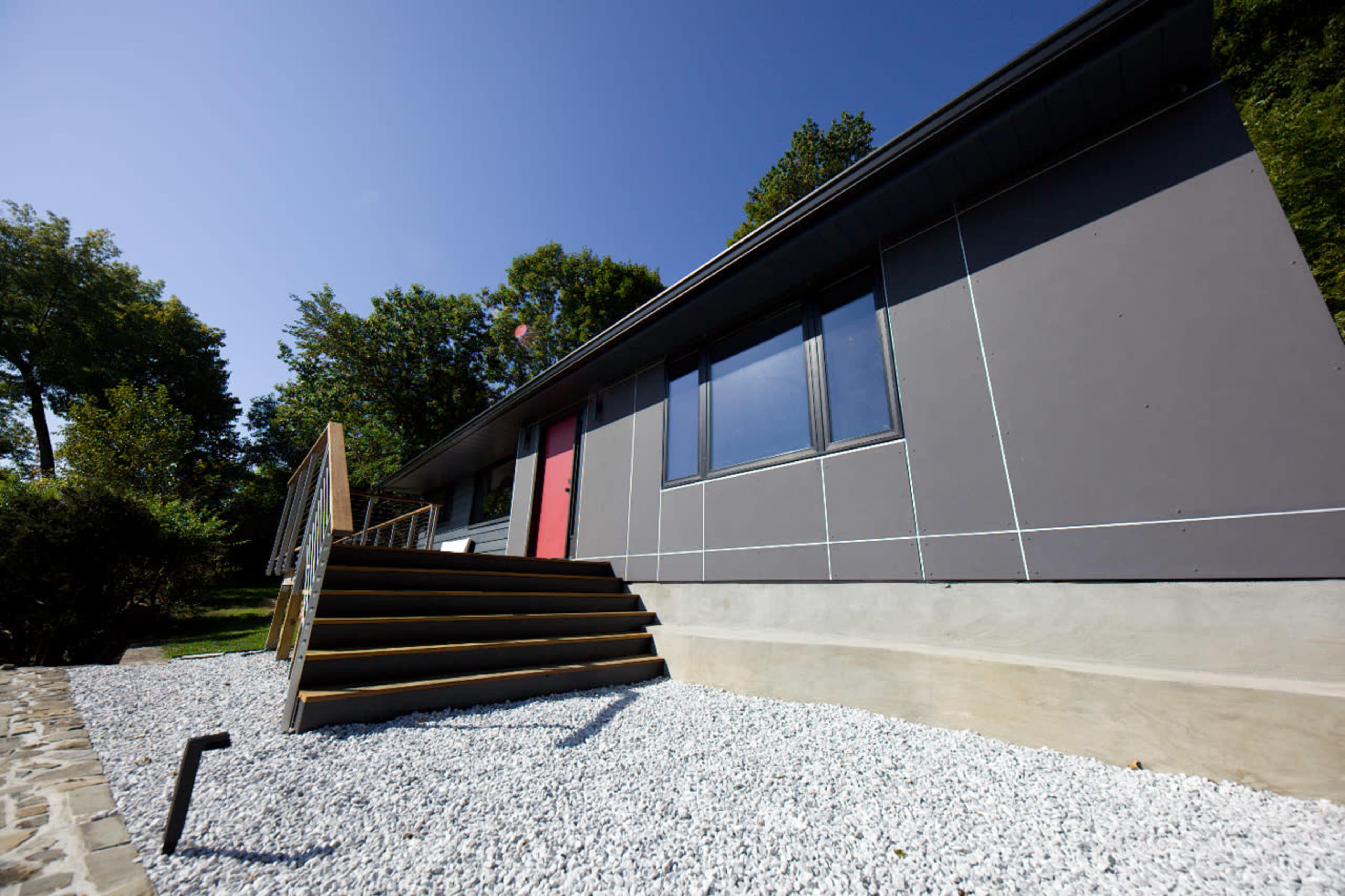 A modern house with a gray exterior, a distinctive red door, and a set of wooden stairs leading to a landscaped gravel area.