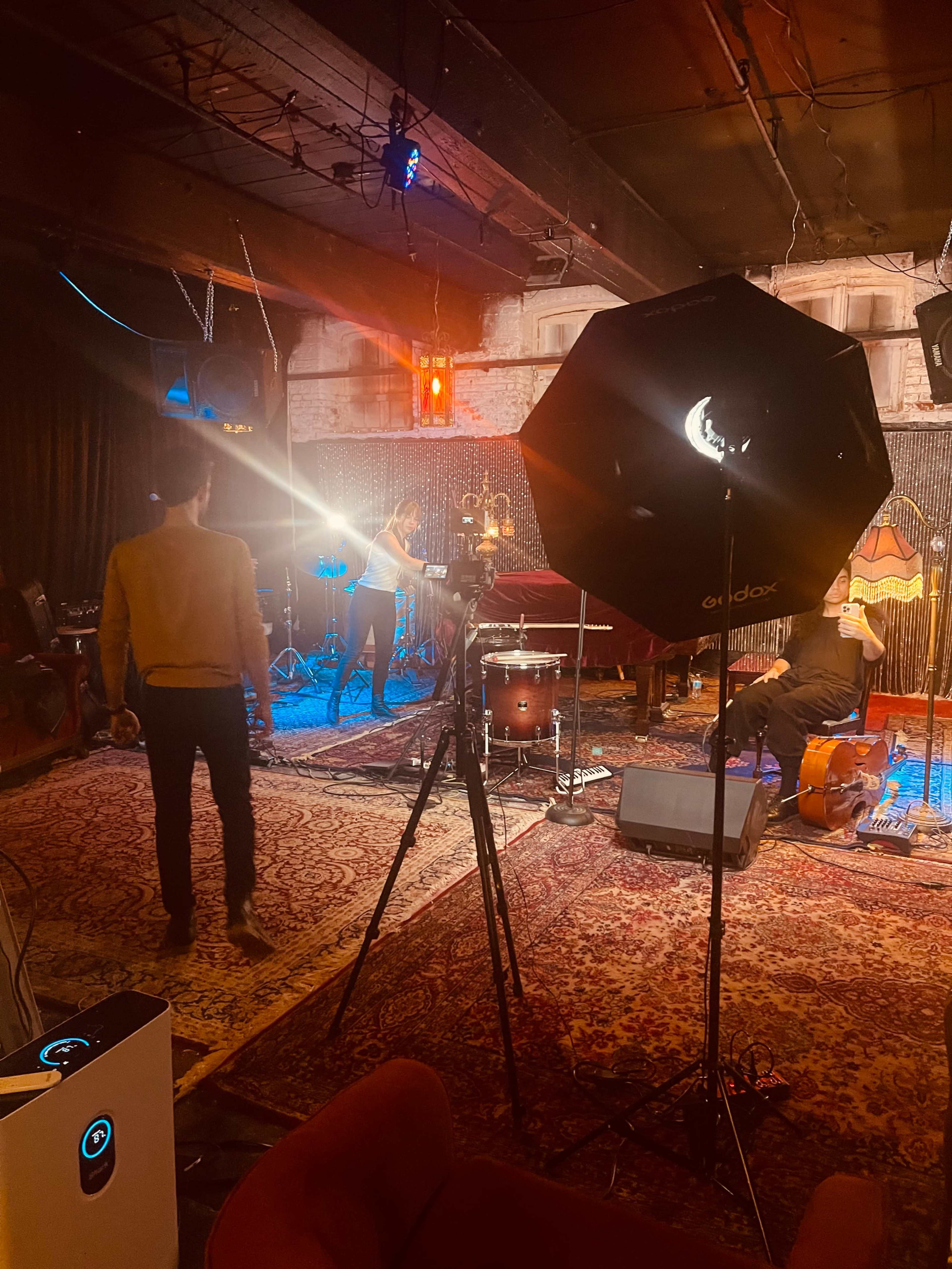 A musician plays guitar on stage while a camera setup captures the performance in a dimly lit venue.