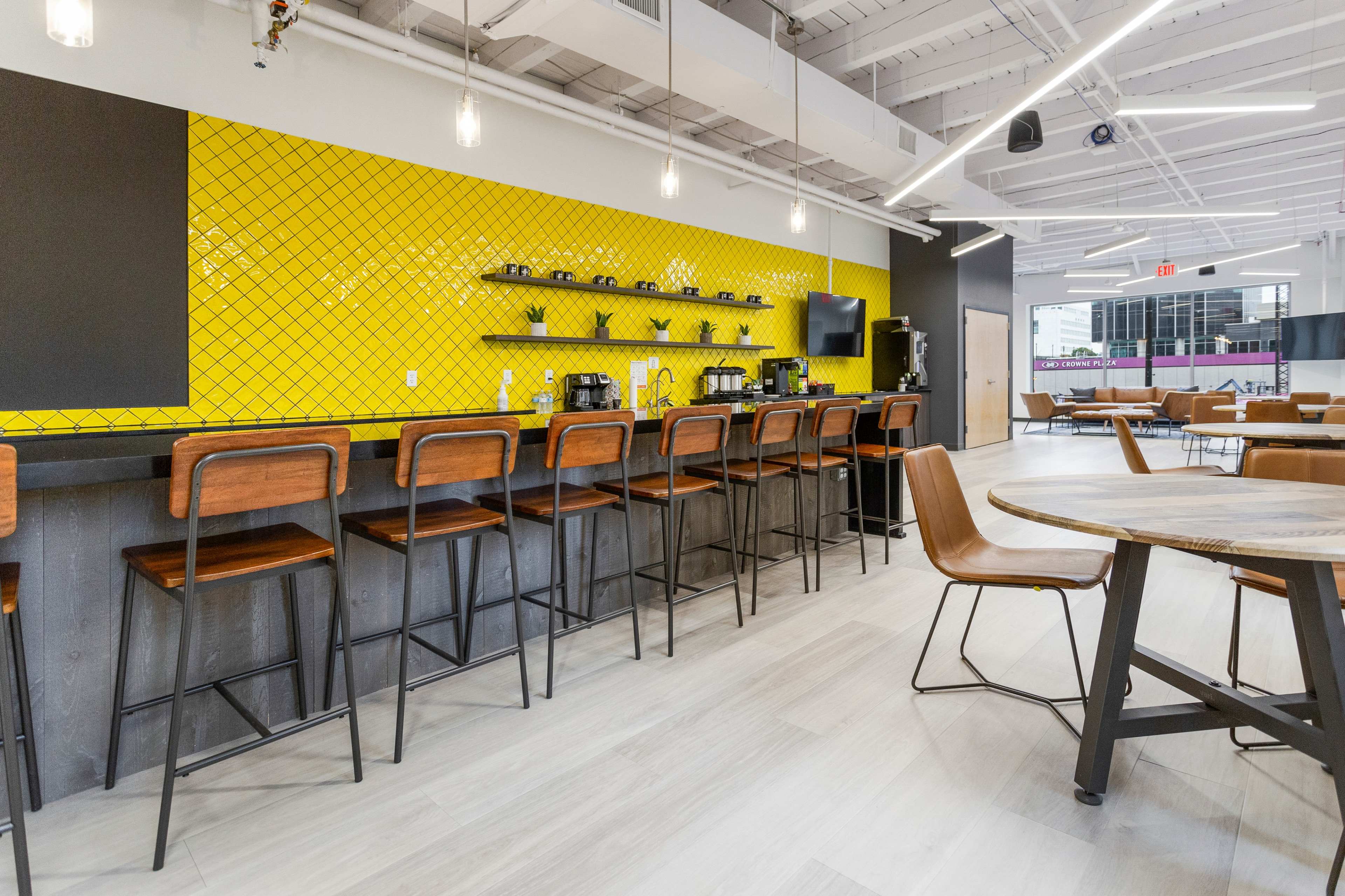 A modern kitchen space features a long bar with wooden stools, yellow tiled walls, and coffee equipment against a sleek backdrop.