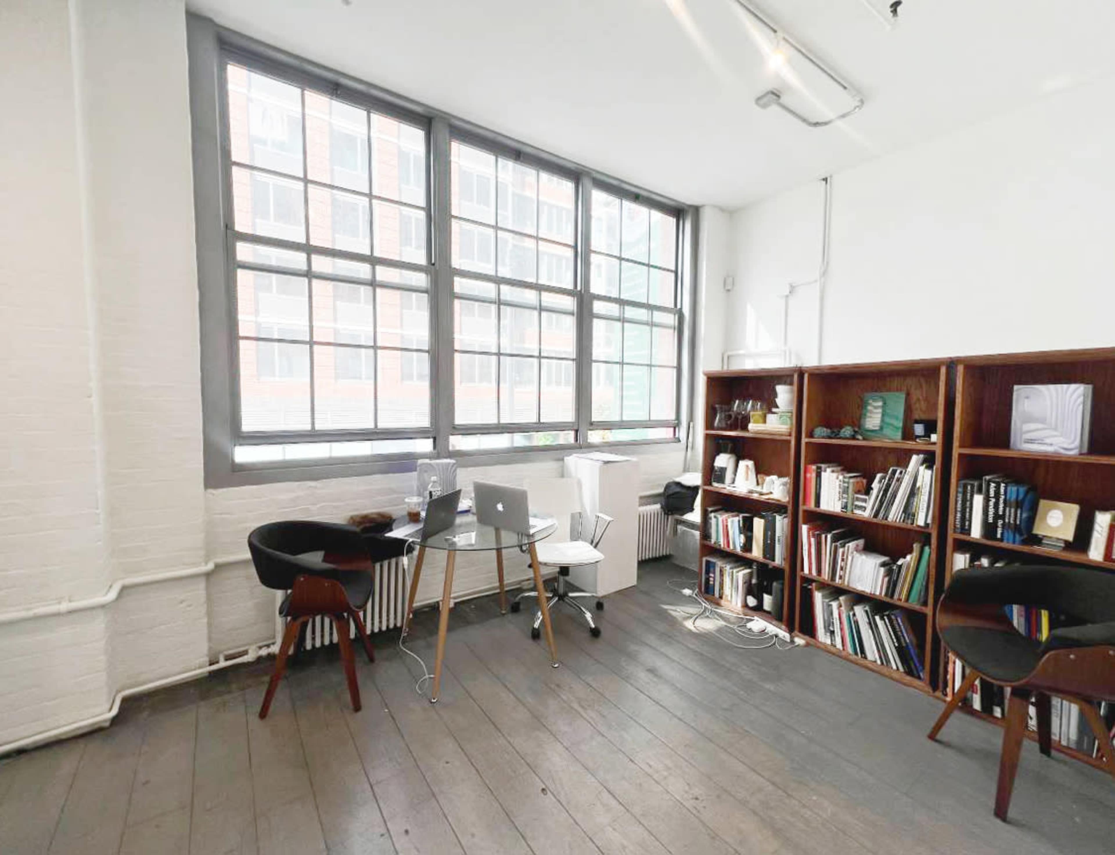 The image shows a bright office space with large windows, a glass table, two chairs, and a bookshelf filled with books.