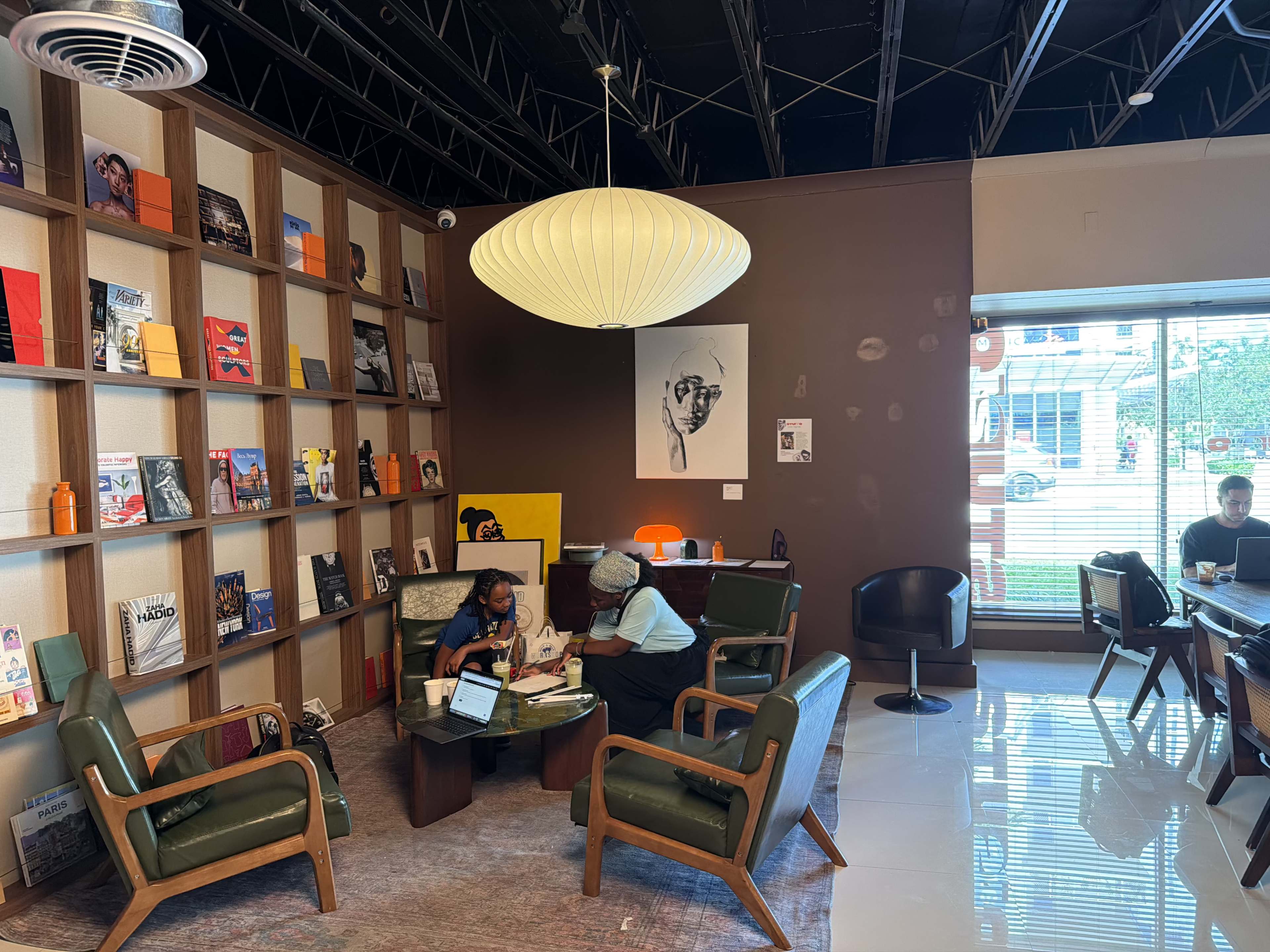 The image shows a modern coffee shop interior with wooden shelves displaying books and magazines, several patrons seated in comfortable chairs, and large windows allowing natural light to enter.