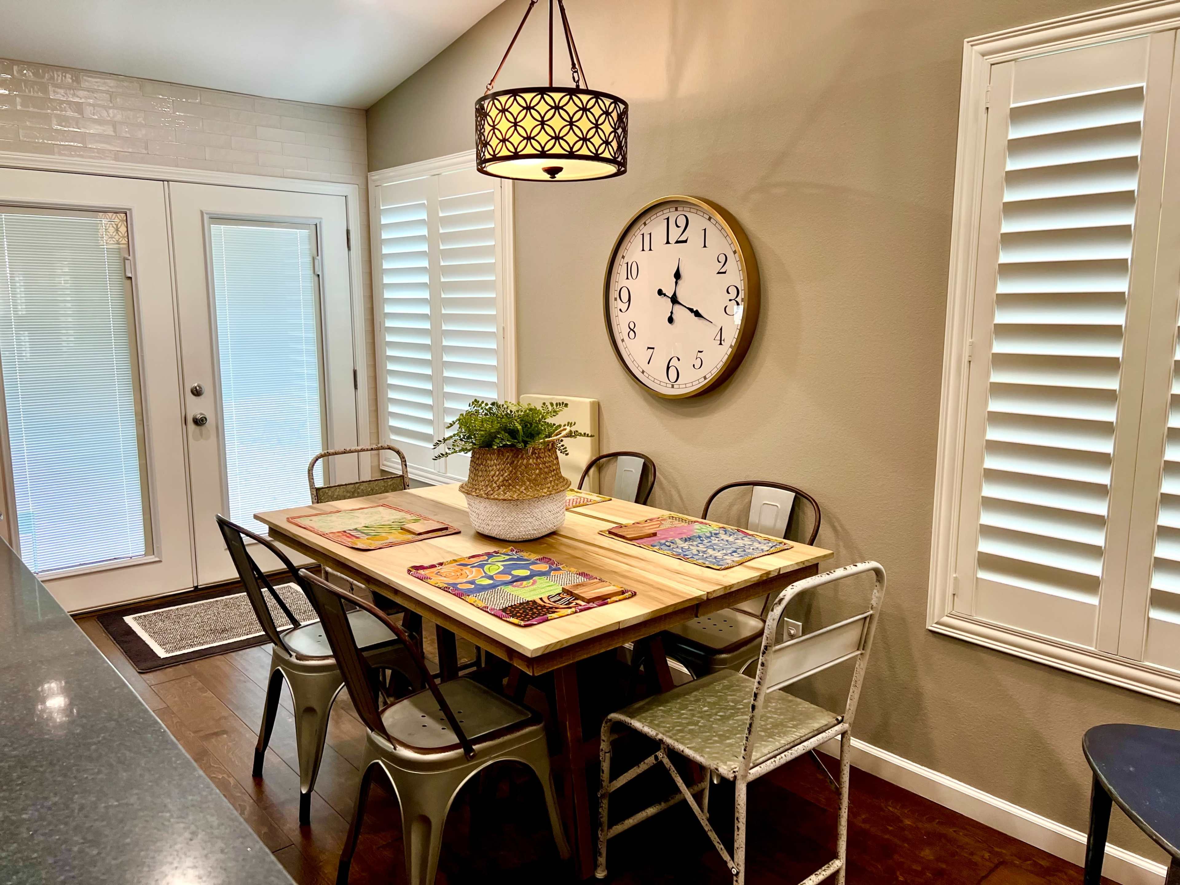 A dining area features a wooden table surrounded by metal chairs, with a wall clock and shuttered windows in the background.