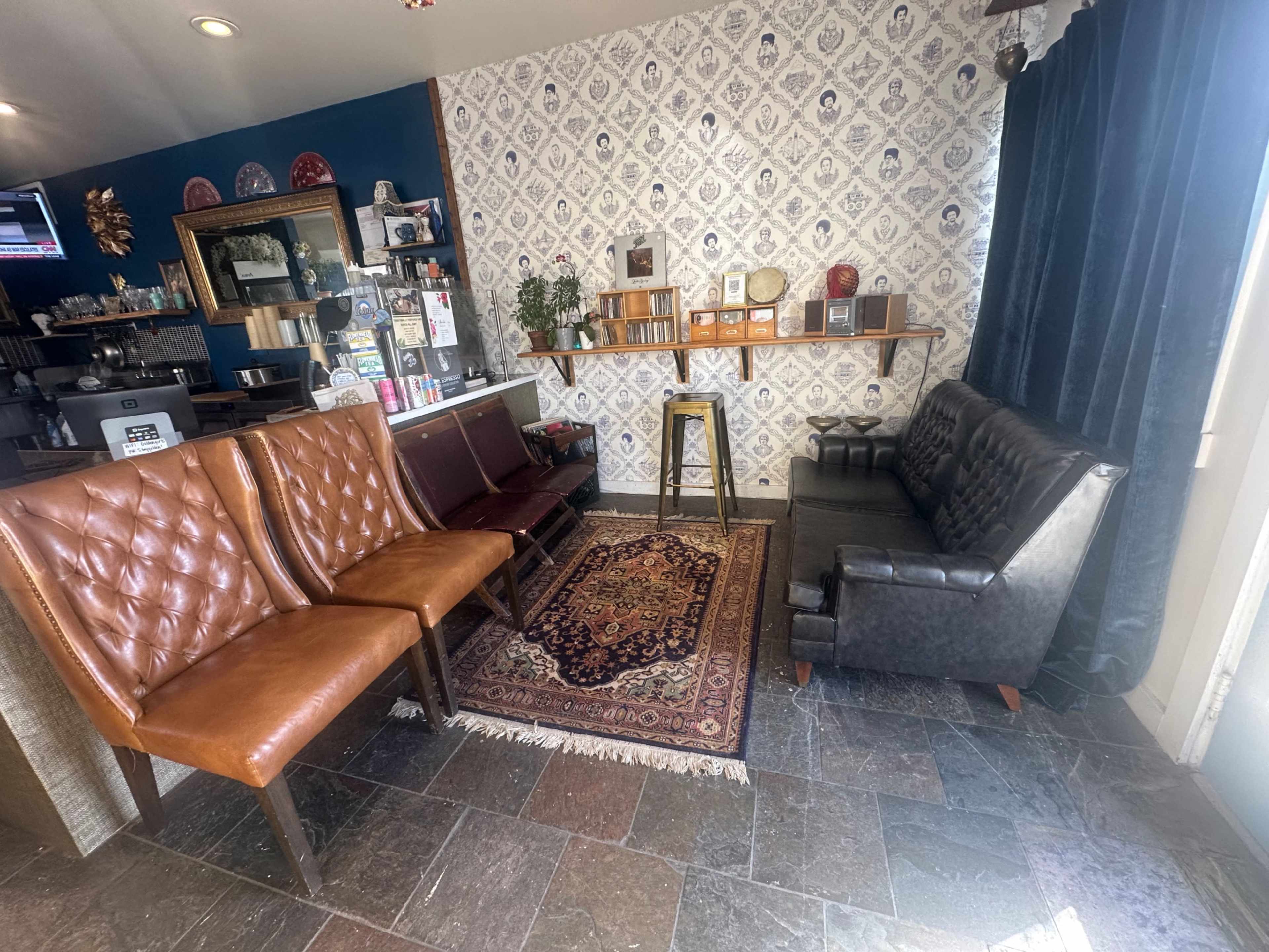 The image shows a cozy interior space featuring two wooden chairs with leather cushions, a dark leather sofa, a patterned area rug, and a decorative wall with shelves holding various items.