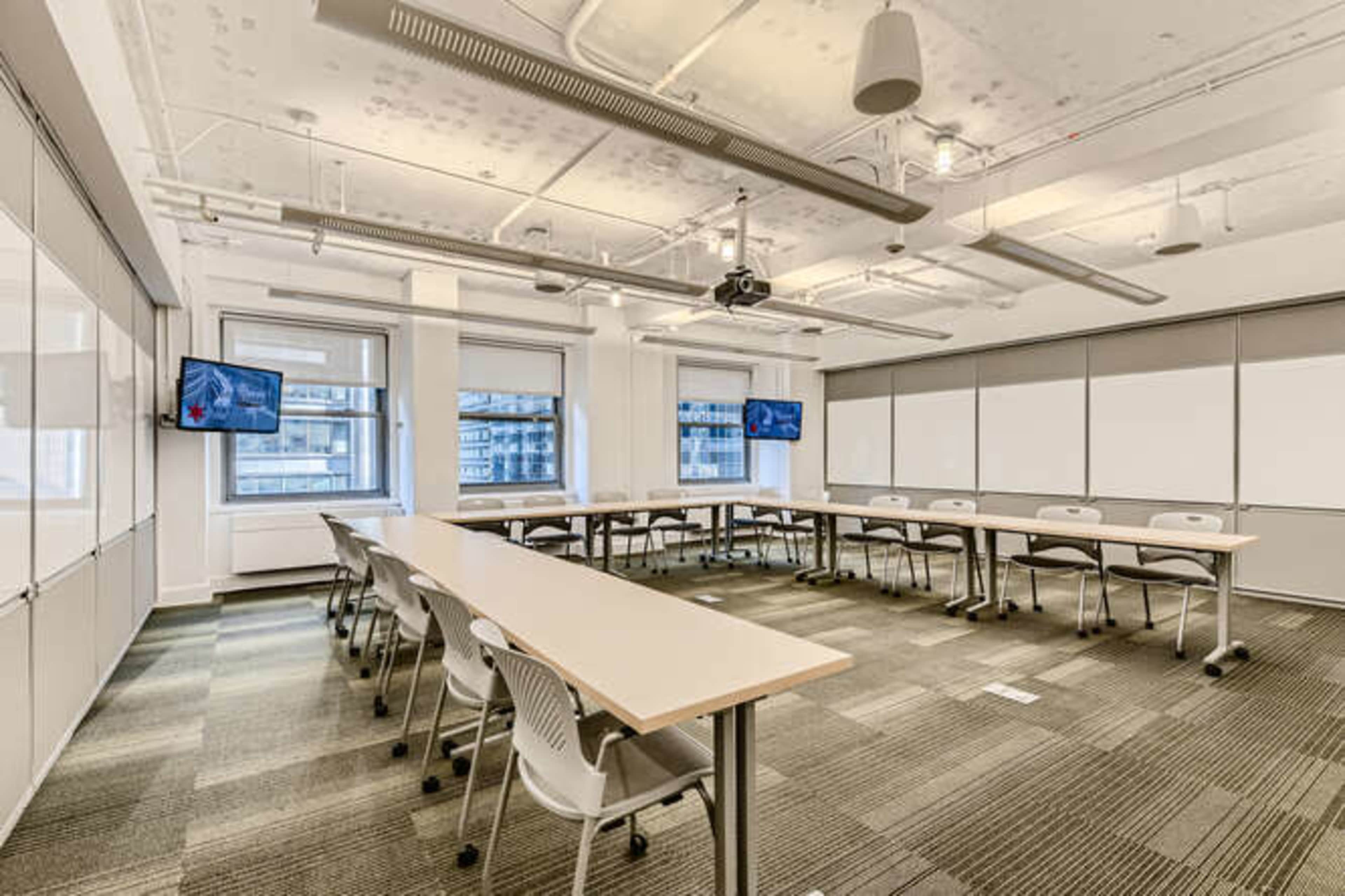 A modern conference room features long tables arranged in a U-shape, with screens mounted on opposite walls and ample overhead lighting.