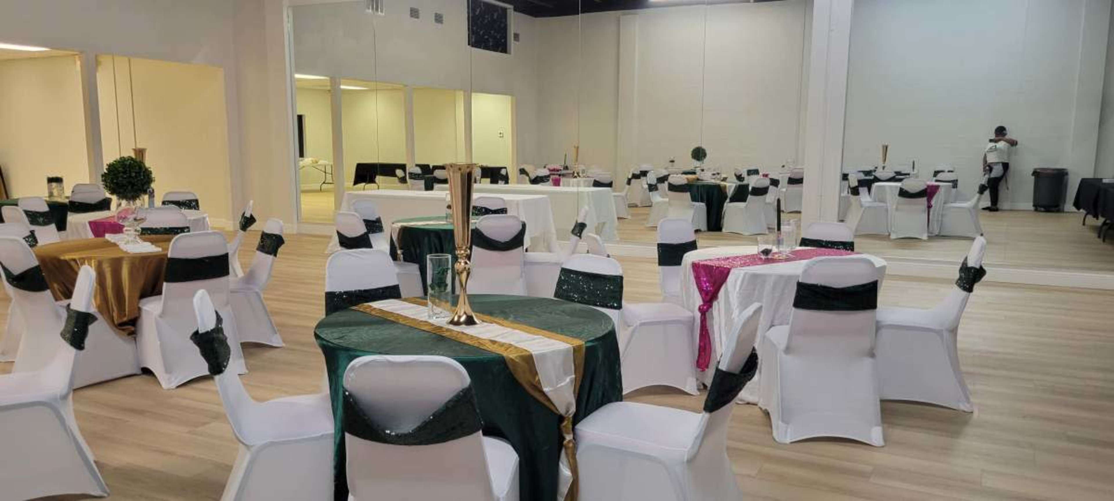 A banquet hall setup with round tables covered in colored cloths and white chairs, some adorned with sashes, and large mirrors on one wall.