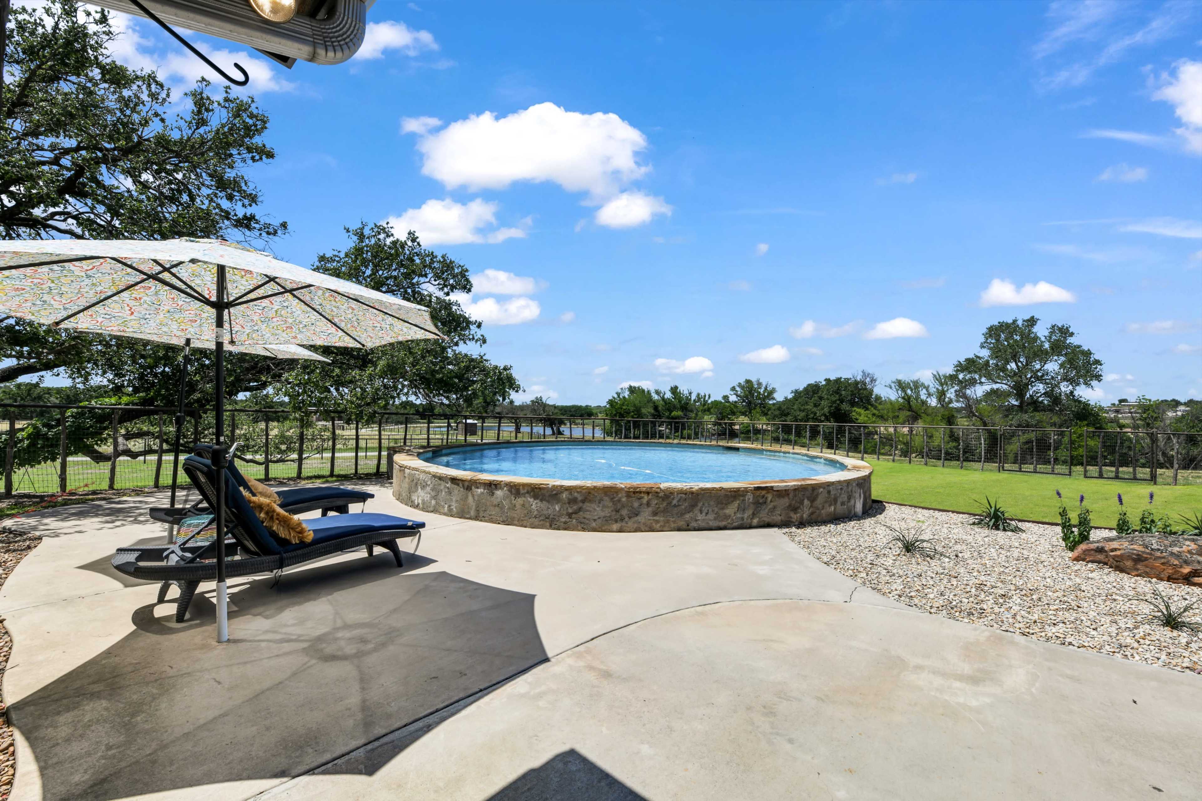 A circular pool with a stone edge is surrounded by a concrete deck and a shaded lounge area featuring a sun umbrella.