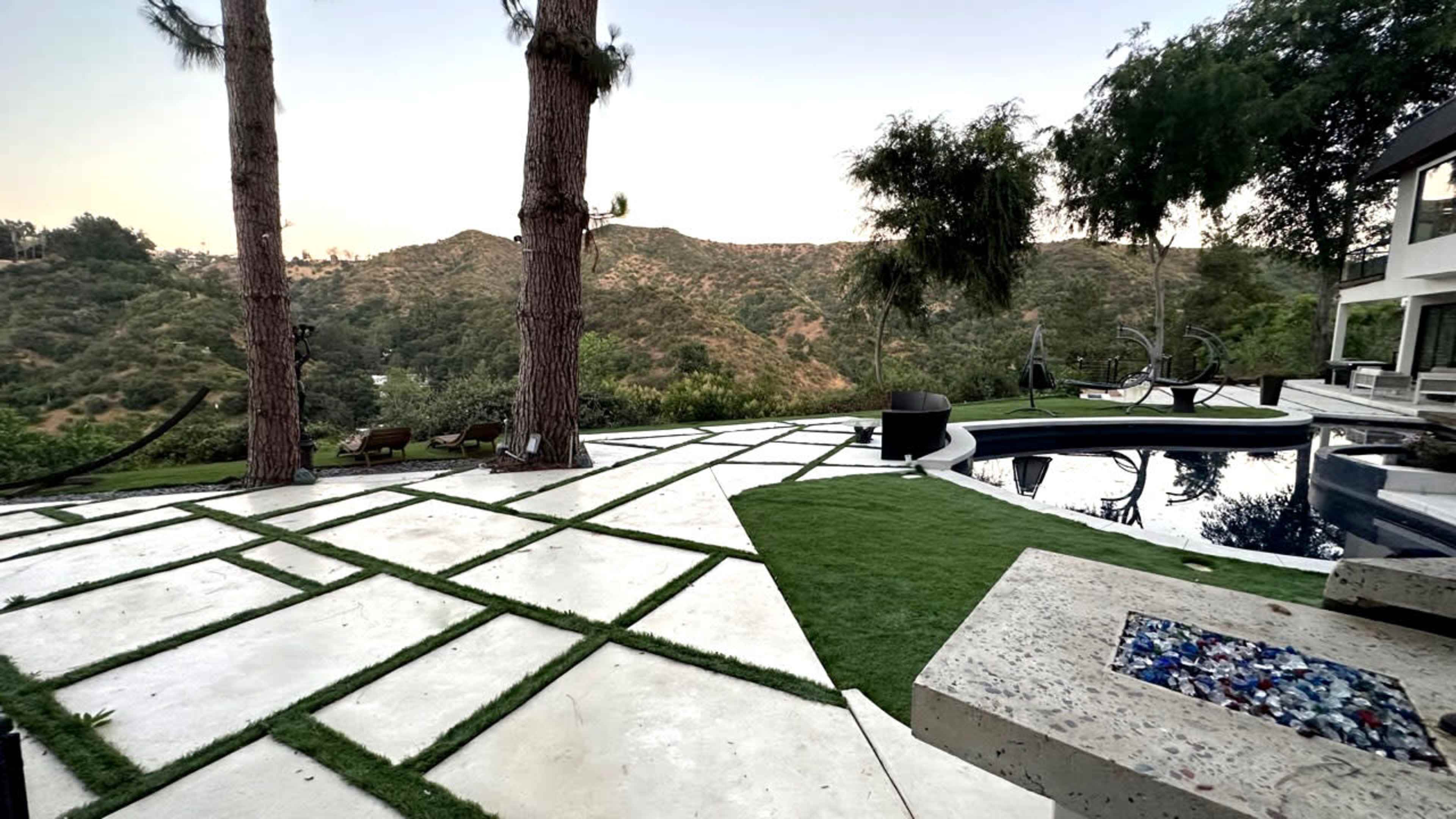A modern outdoor space with a swimming pool, stone patio, and landscaped greenery, set against a hillside backdrop.
