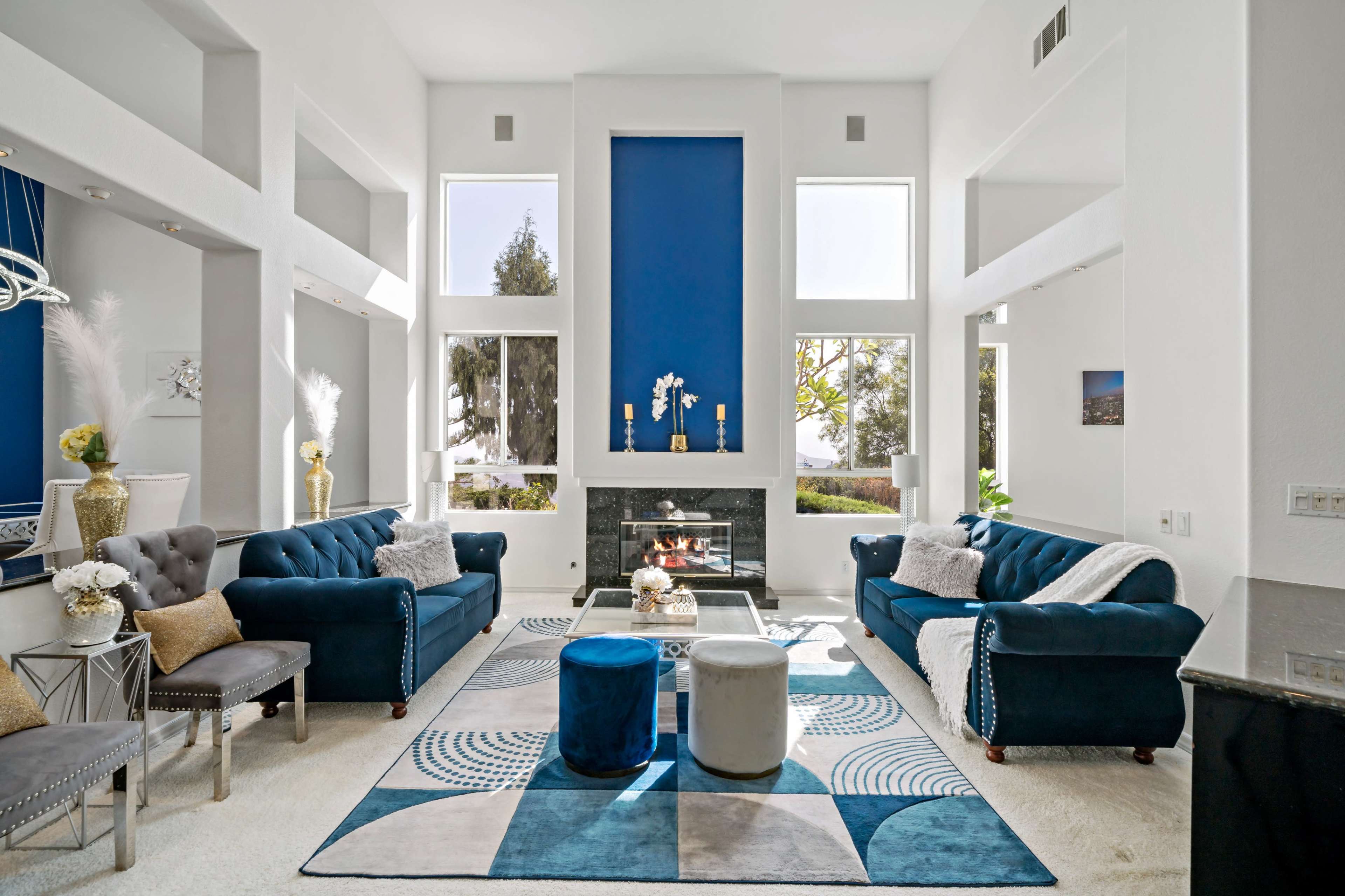 The living room features two blue sofas facing a fireplace, with large windows allowing natural light to fill the space and a geometric rug on the floor.