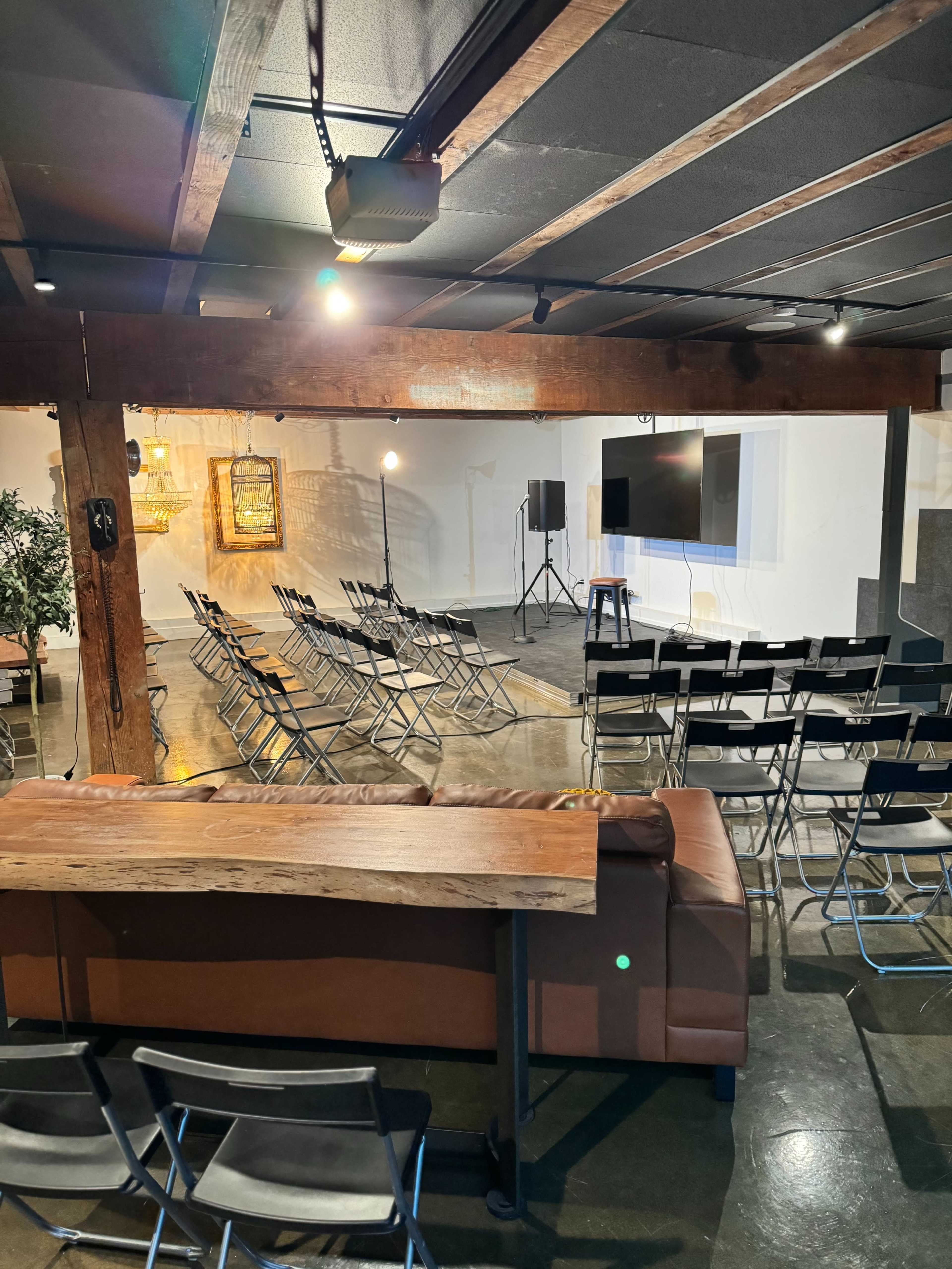 A spacious room with rows of folding chairs arranged facing a screen, accompanied by a couch made of leather and exposed wooden beams.