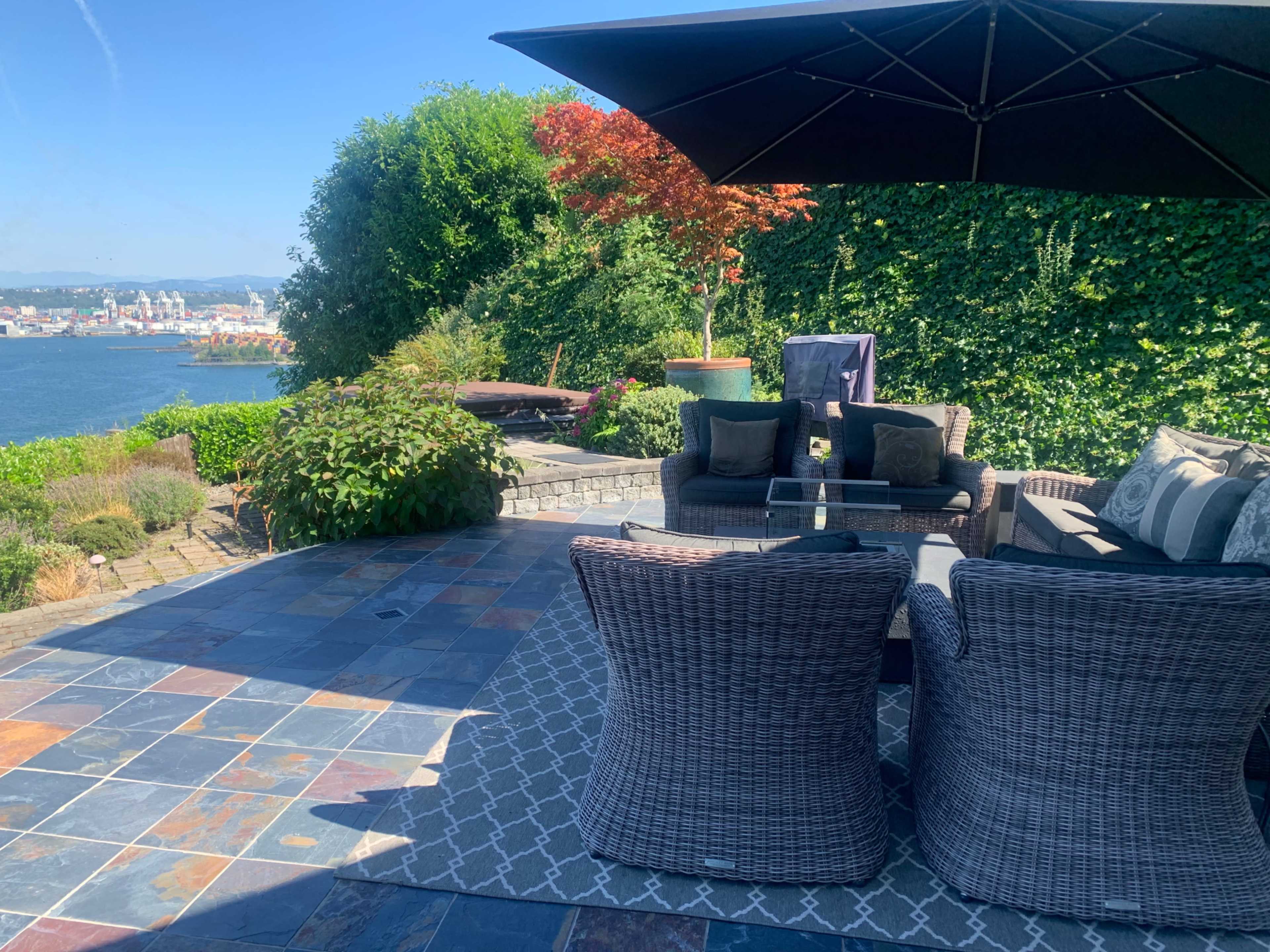 A patio area with wicker seating and a large umbrella, overlooking a river and cityscape in the background.