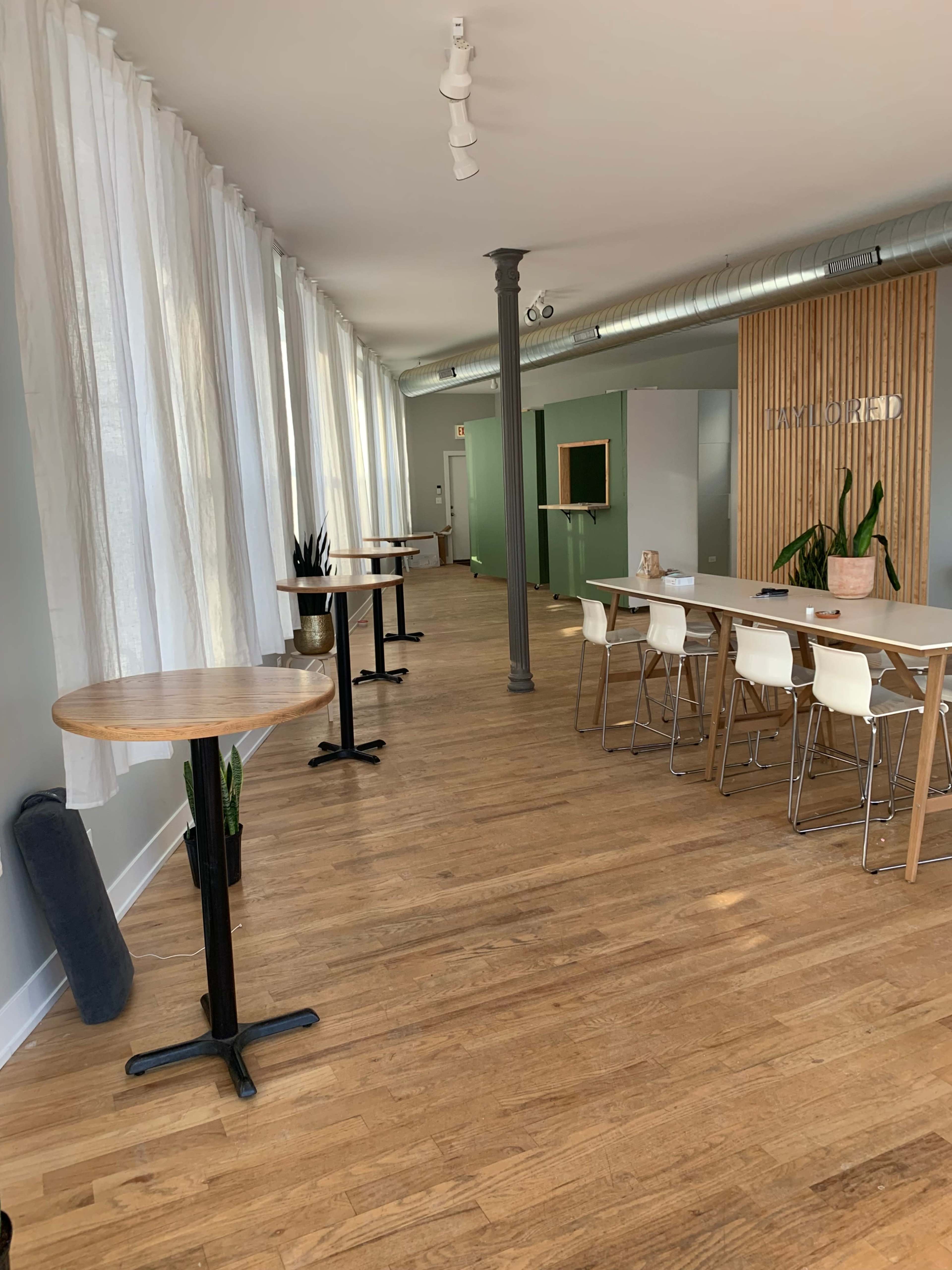 A spacious, well-lit room with large windows, featuring wooden flooring, a mix of small and long tables, and potted plants along the walls.