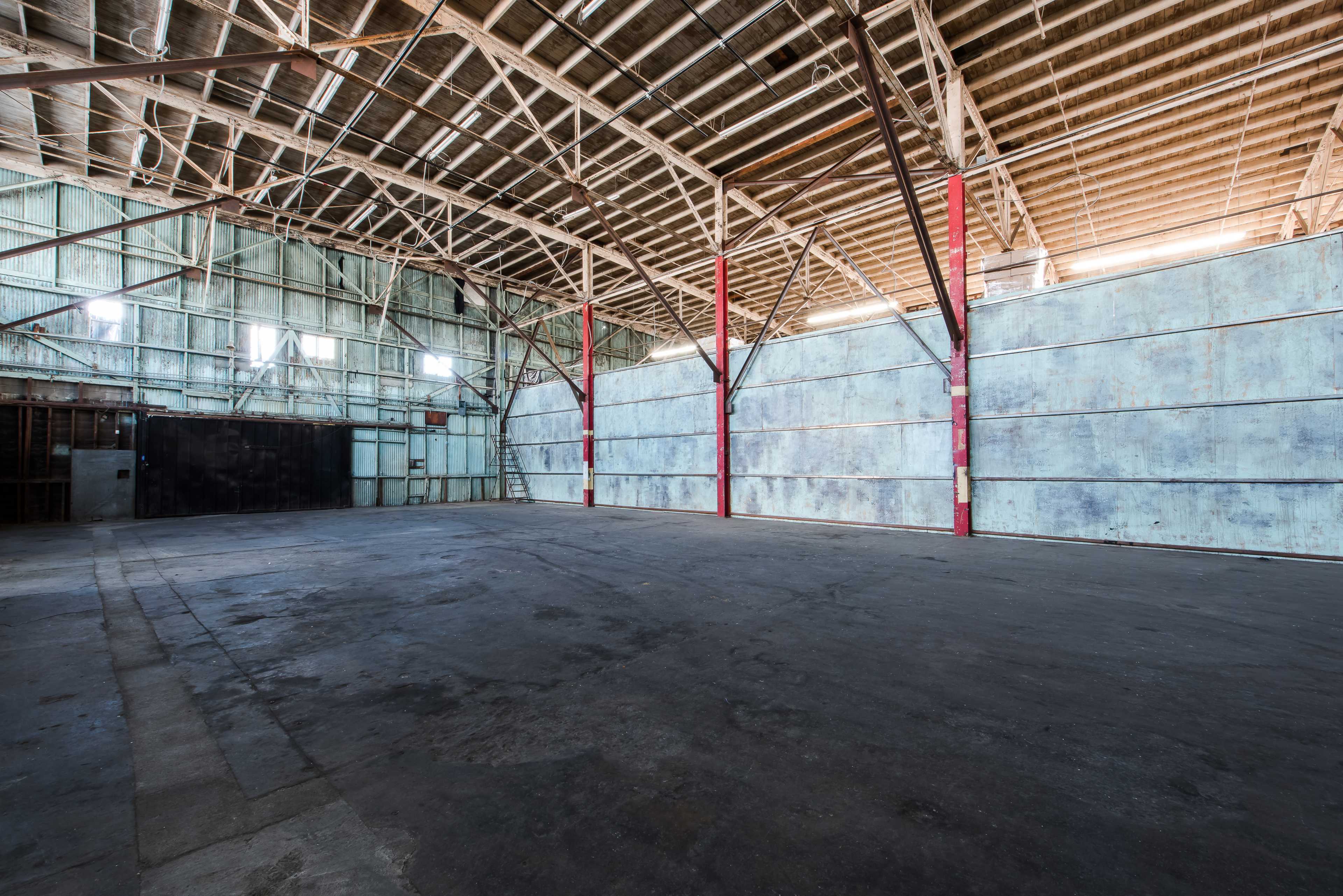 The image shows a large, empty warehouse with high ceilings, exposed beams, and concrete flooring.
