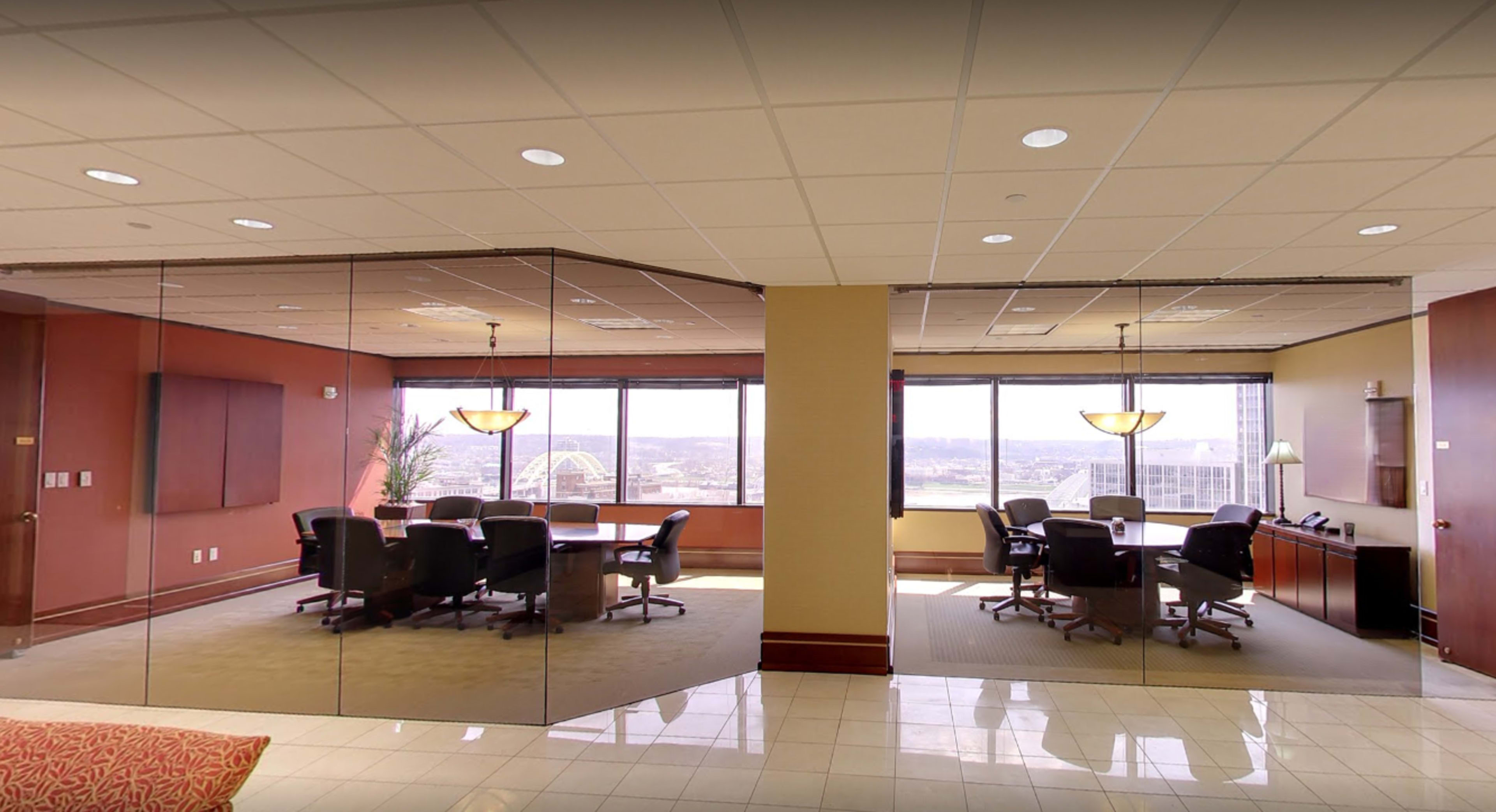 The image shows a modern office space with glass walls separating two conference rooms, both furnished with large tables and chairs, alongside large windows offering a view of the city.