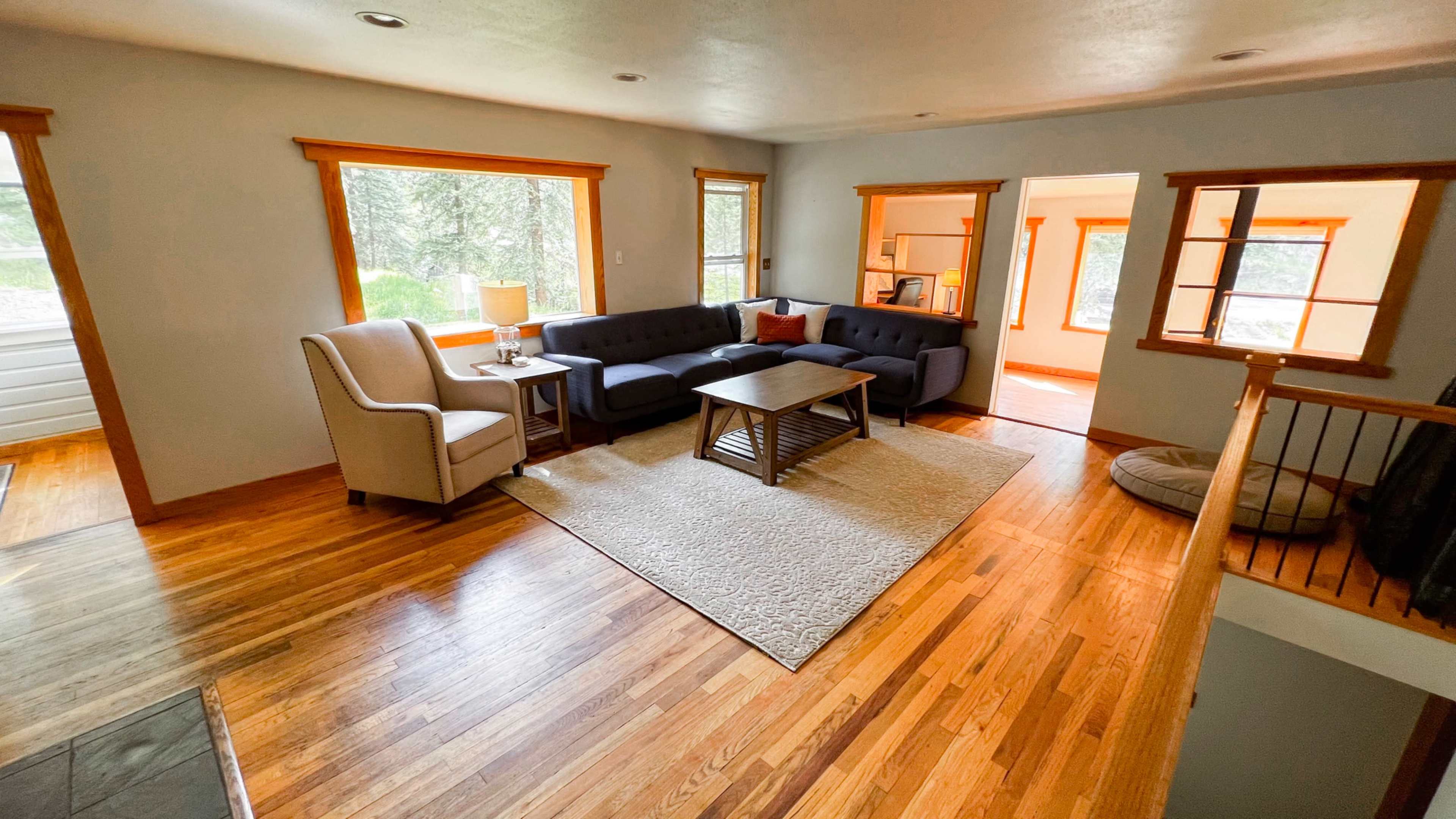 A living room with a sectional sofa, a coffee table, and a light-colored rug, situated near large windows that provide natural light.