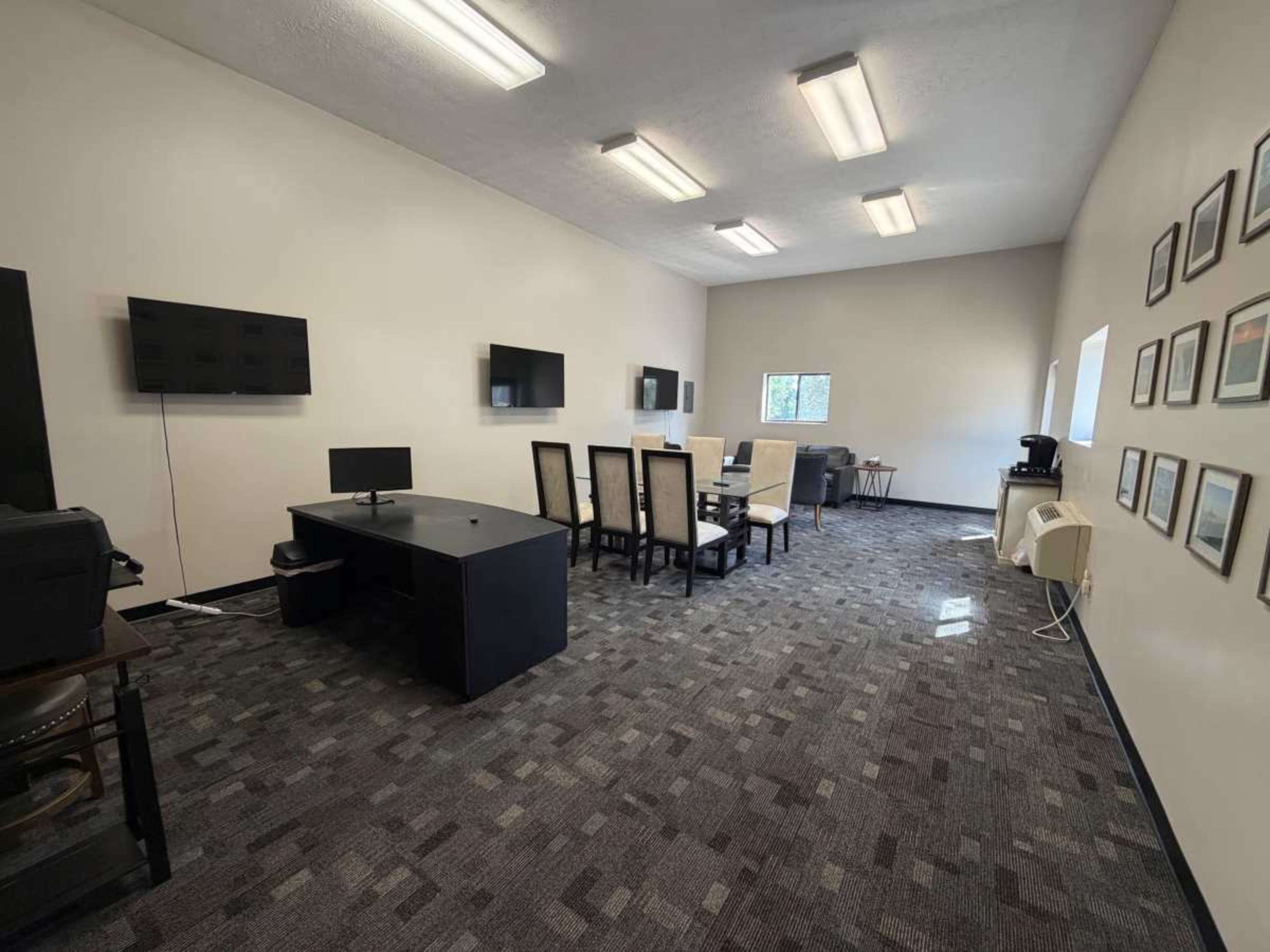 The image shows a spacious office or meeting room featuring desks, chairs, multiple screens mounted on the walls, and framed pictures.