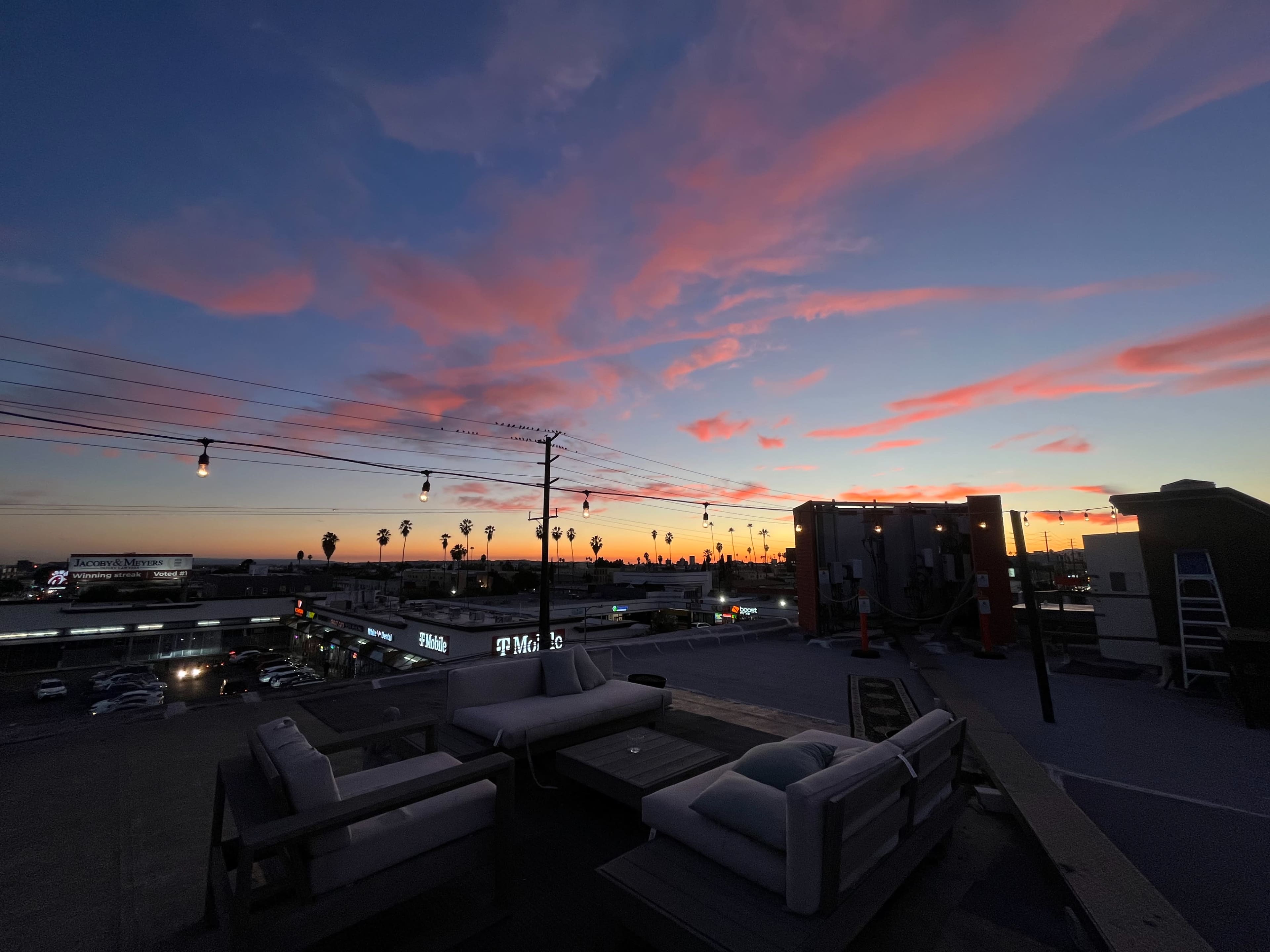 The image shows a rooftop scene at sunset, featuring pink and purple clouds against a darkening sky, with a view of palm trees and city lights in the distance.