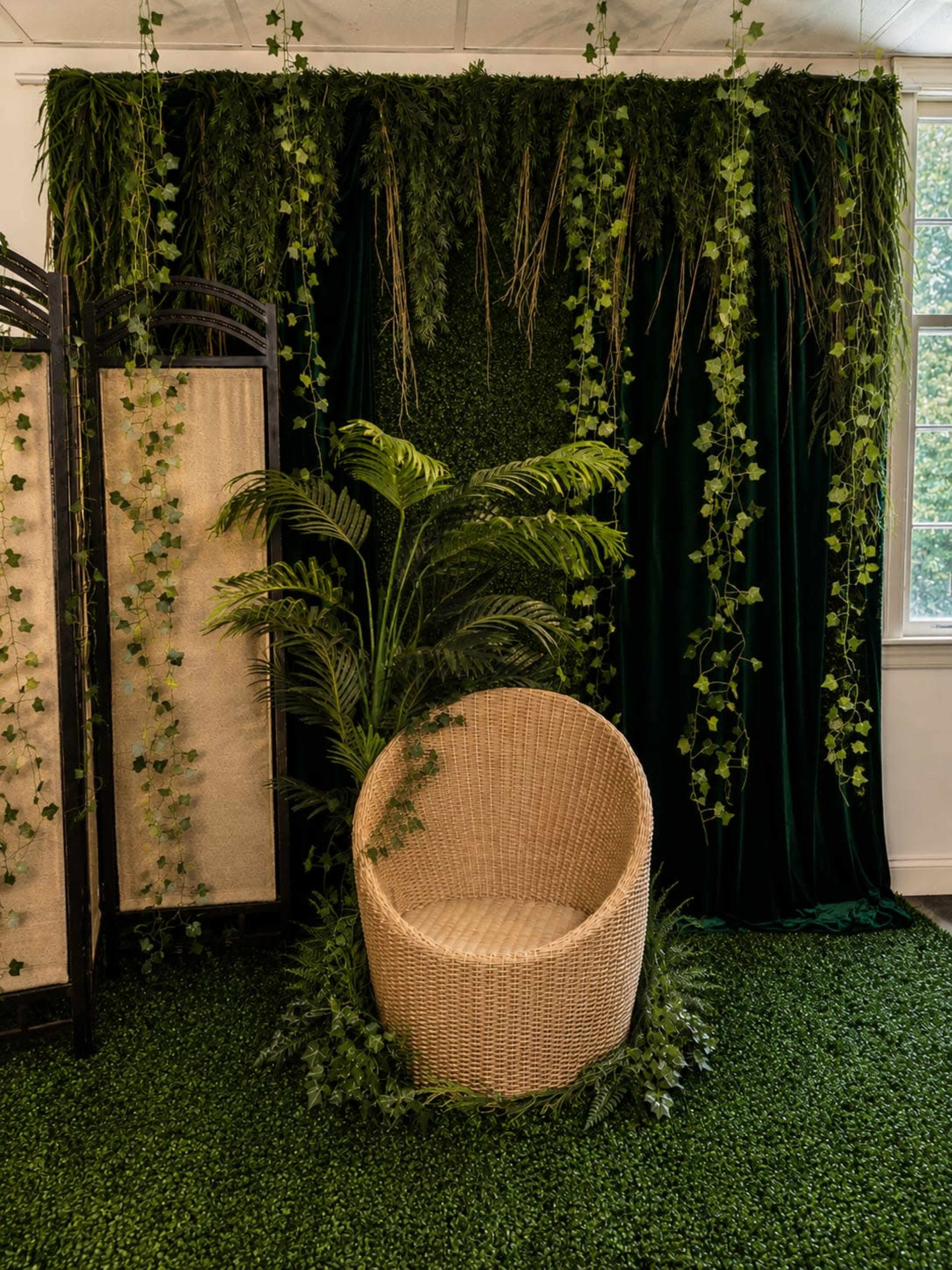The image shows a cozy corner setup with a wicker chair surrounded by artificial greenery and a backdrop of hanging vines and lush fabric.