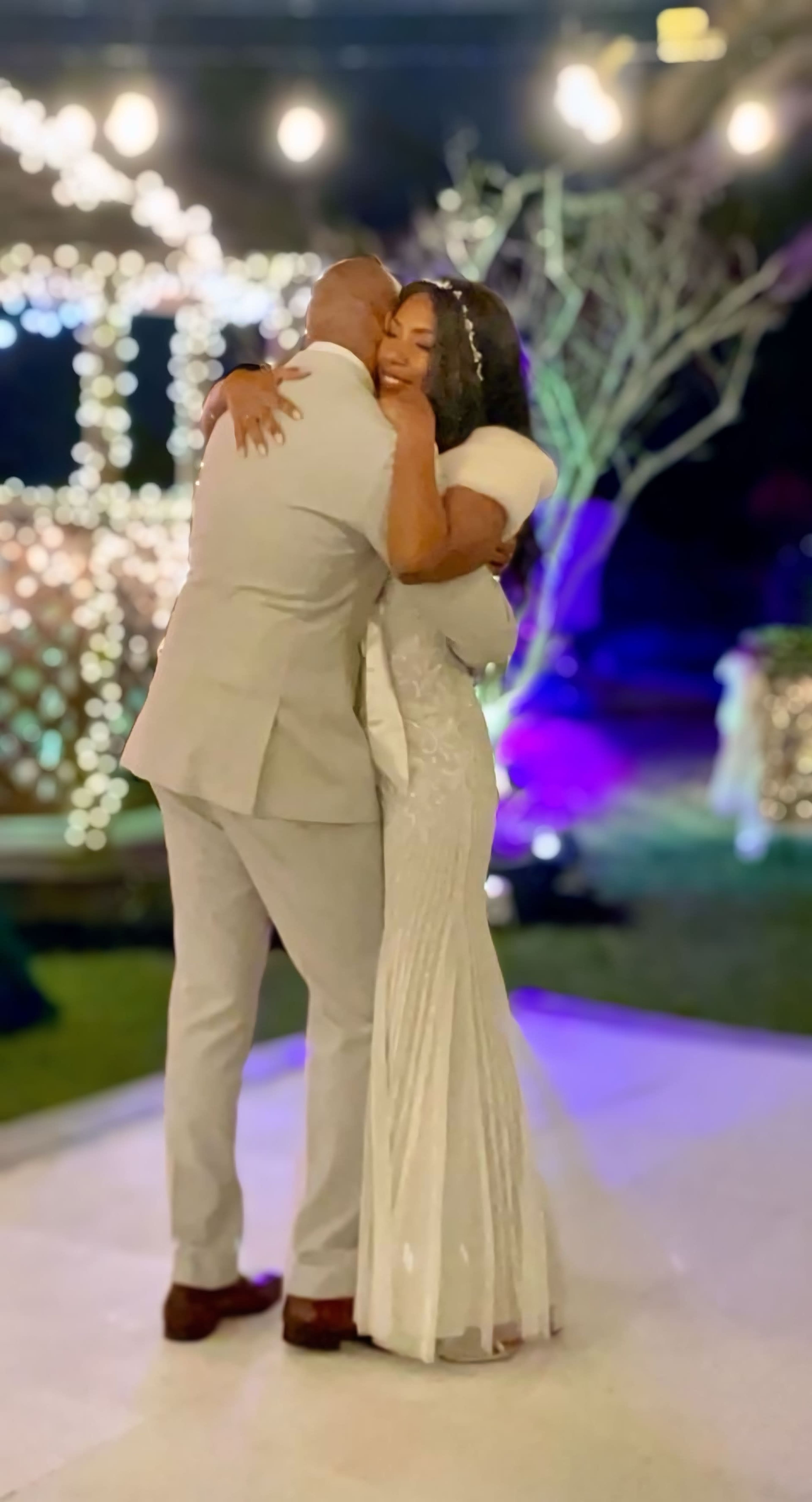 A bride and groom embrace on a decorated outdoor dance floor illuminated by colorful lights.