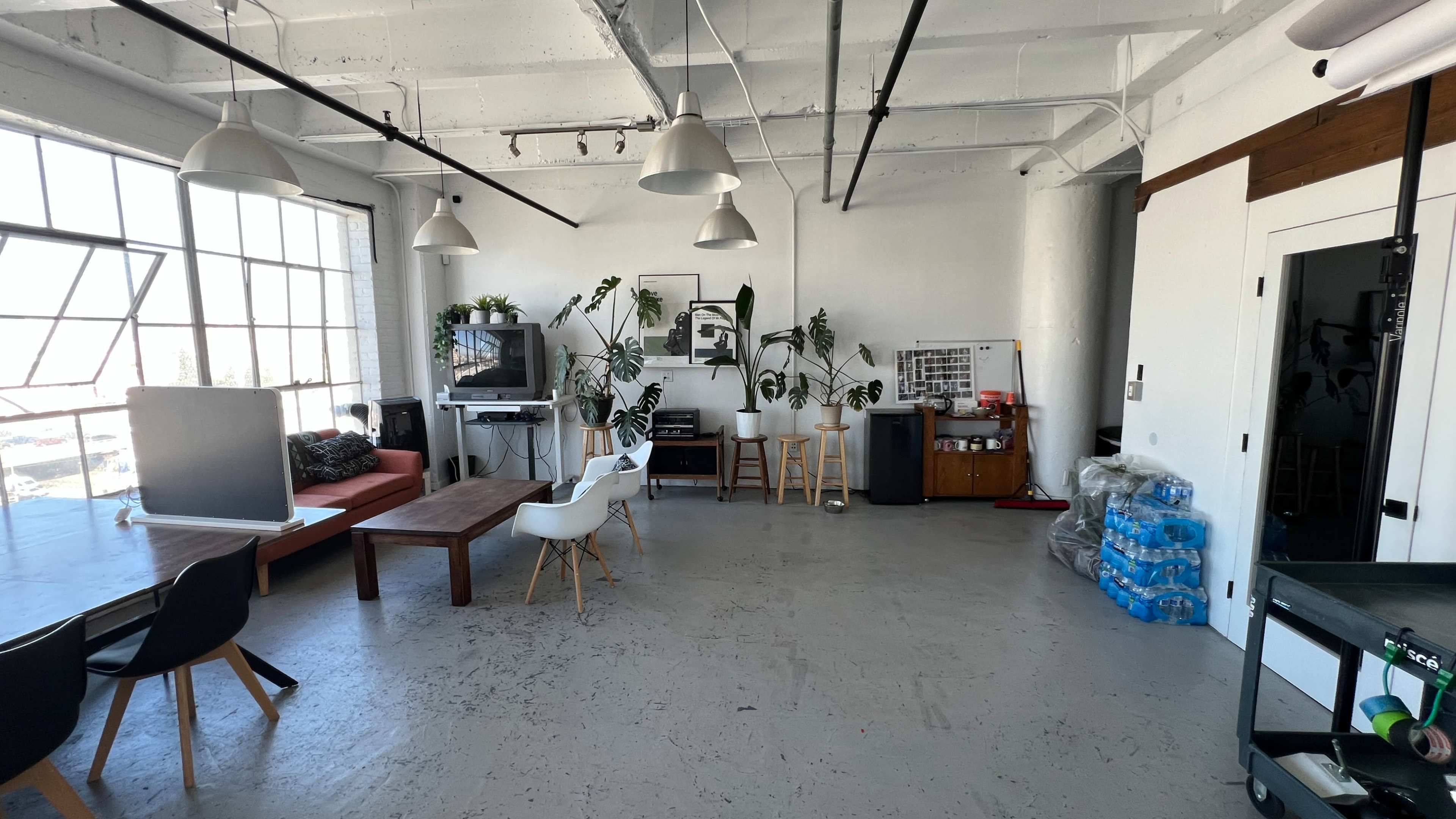 The image shows a spacious office interior with large windows, a seating area, a dining table, and various plants.