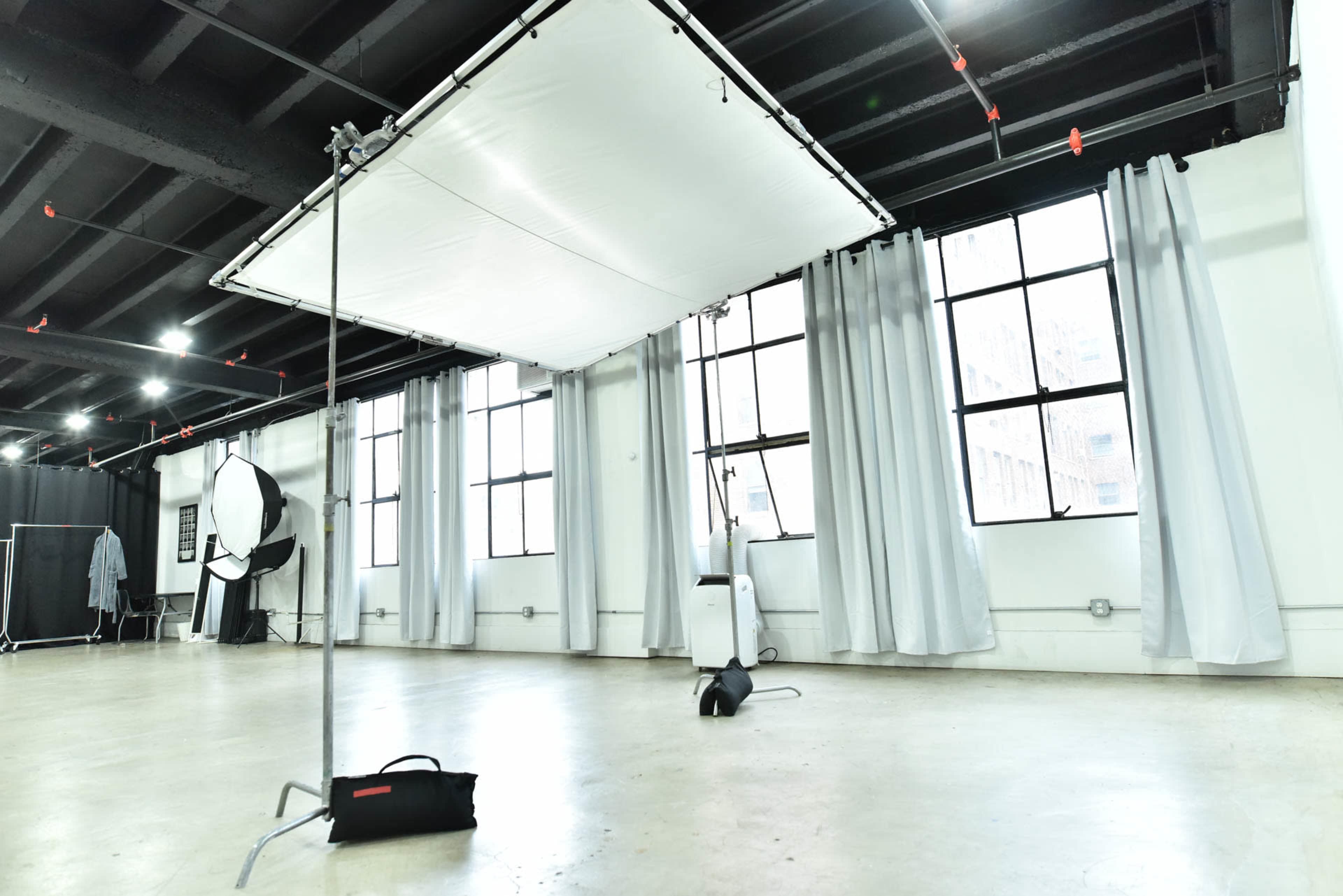 The image shows a spacious photography studio with large windows, gray curtains, and lighting equipment set up for a shoot.