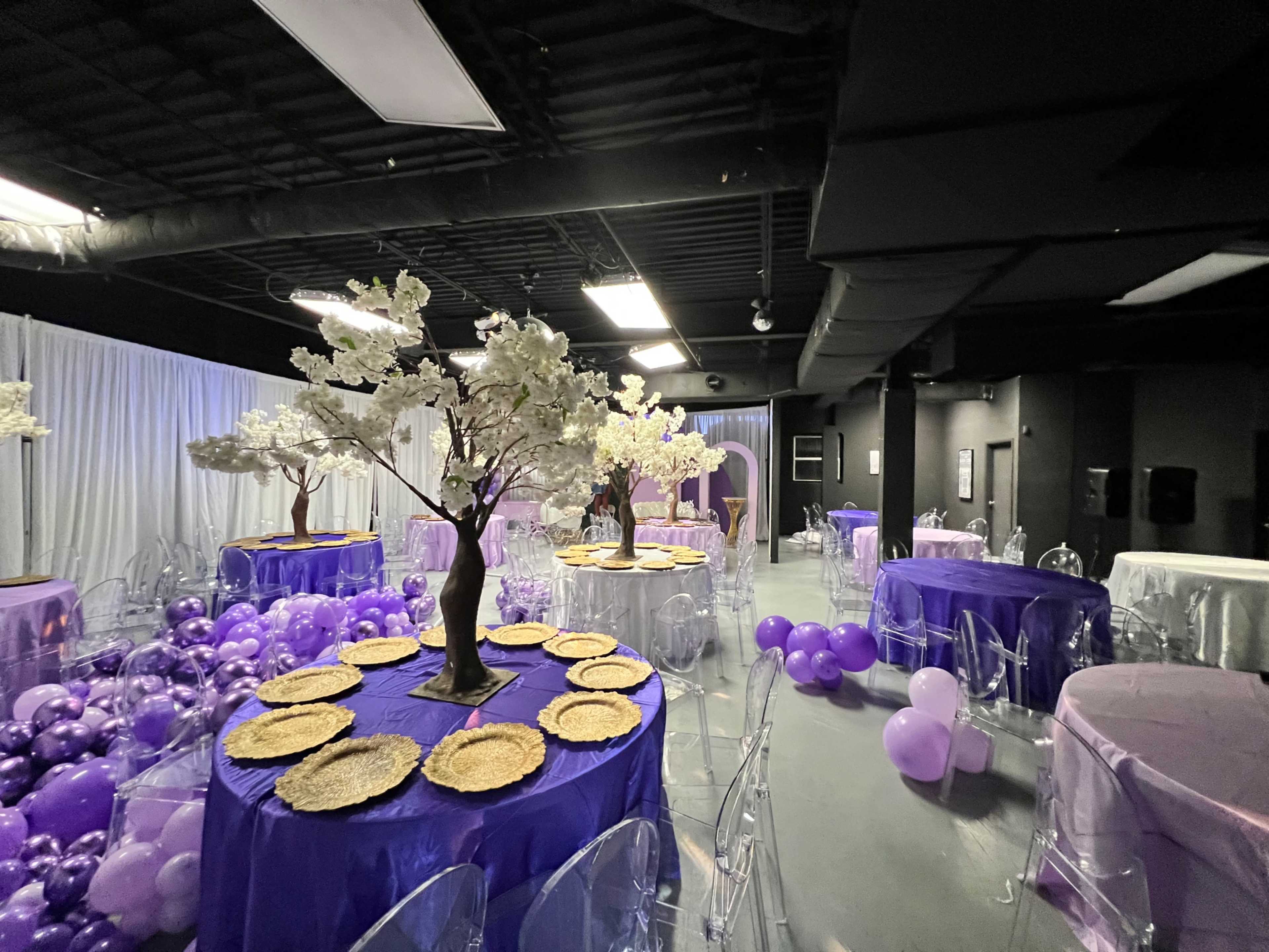 An event space with round tables covered in purple tablecloths, decorated with floral centerpieces and surrounded by clusters of purple balloons.
