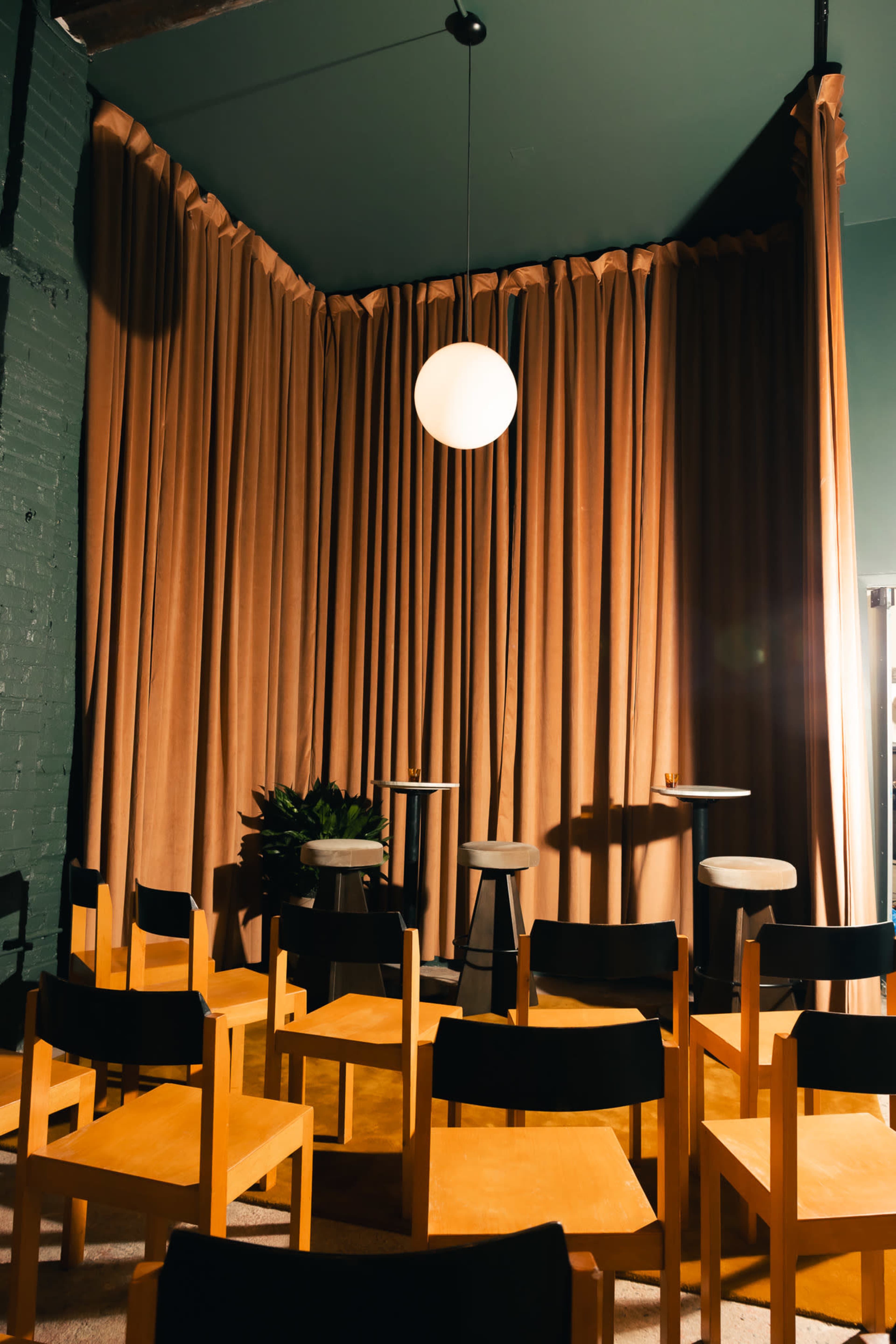 The image shows a small performance space with wooden chairs arranged in front of a stage area, featuring soft brown curtains and a round pendant light hanging above.