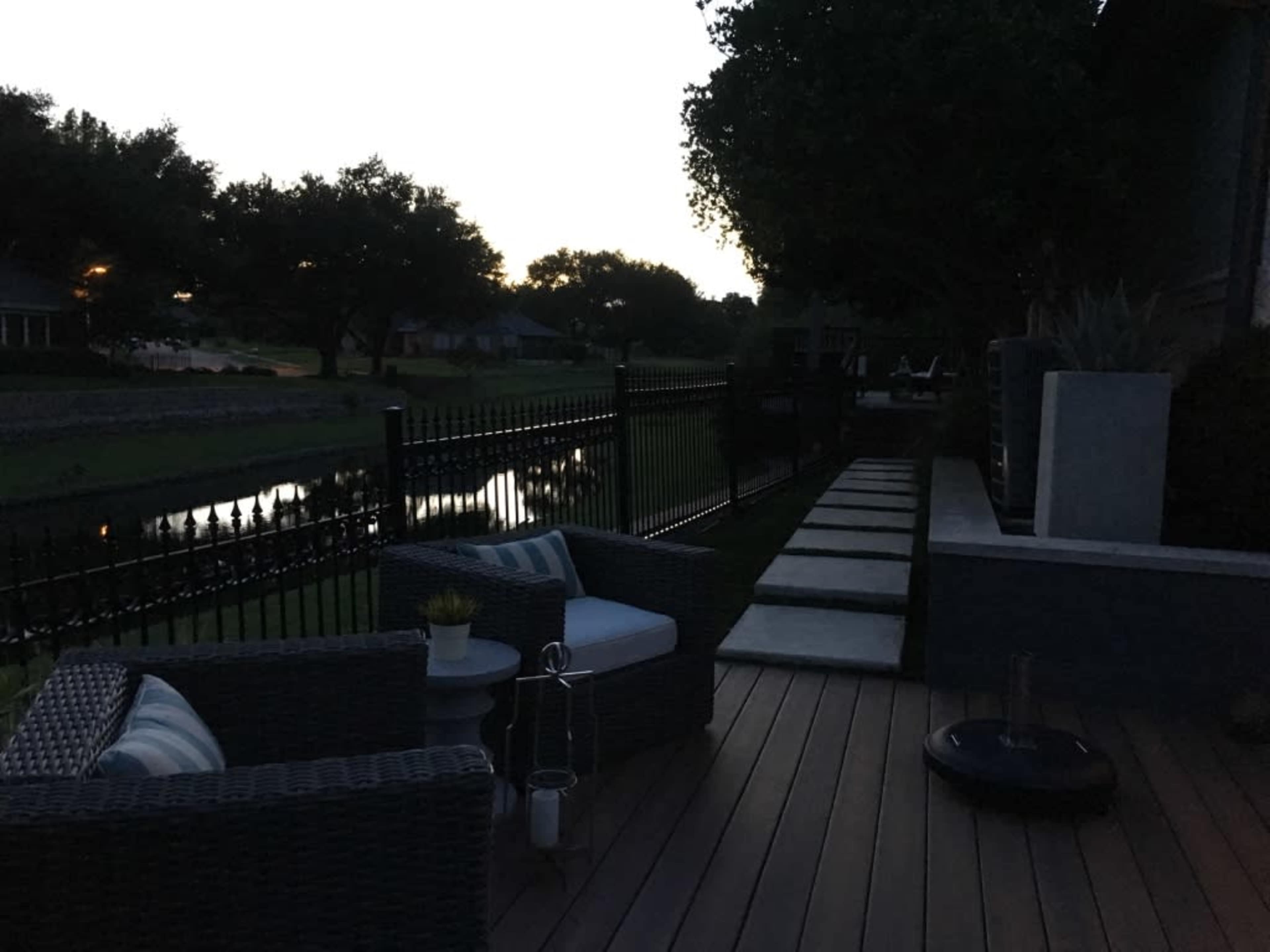 A patio with wicker chairs and stone pavers overlooks a darkened yard and a reflective water feature at dusk.