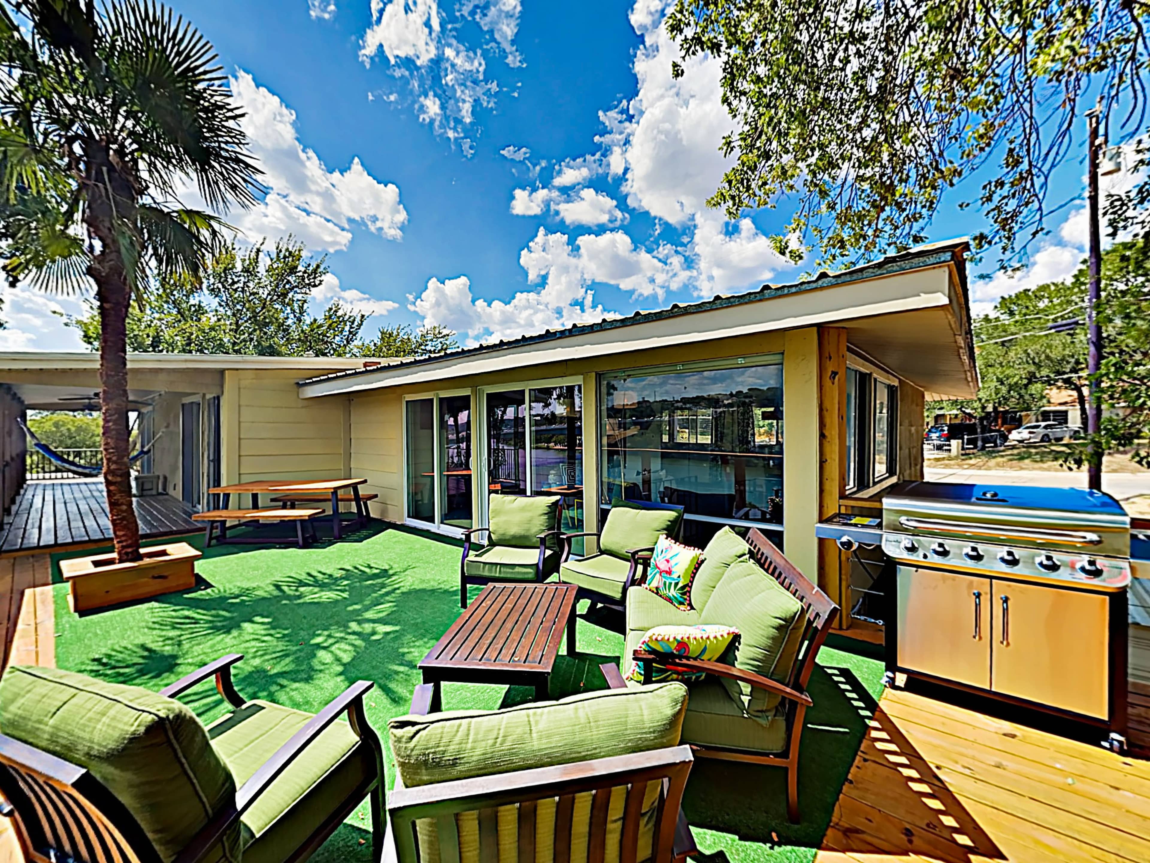 The image shows a patio area with green seating, a gas grill, and a view of trees and a sky filled with clouds.