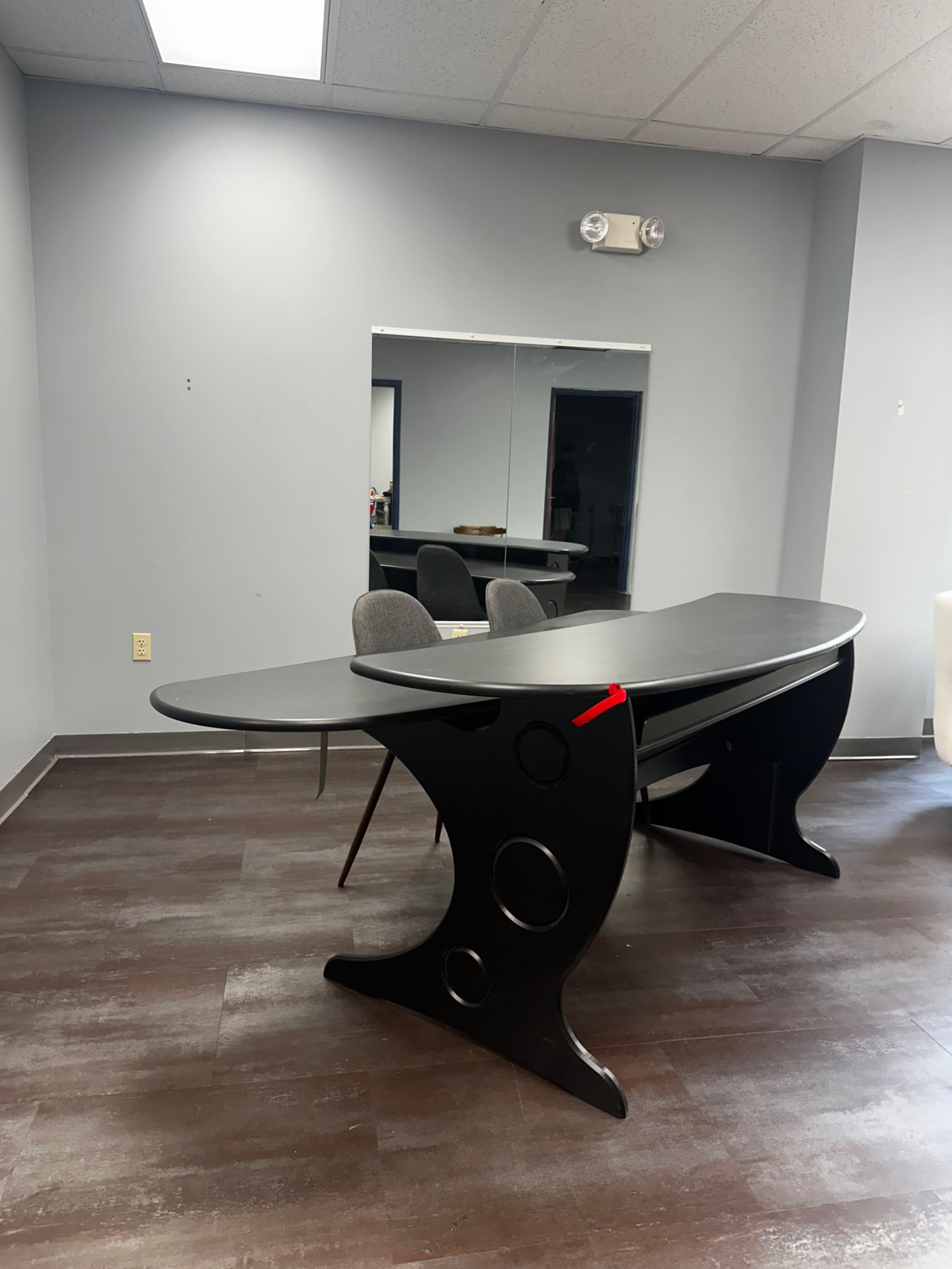 The image shows a modern black desk with a curved design positioned in a room that features gray walls and a large mirror.