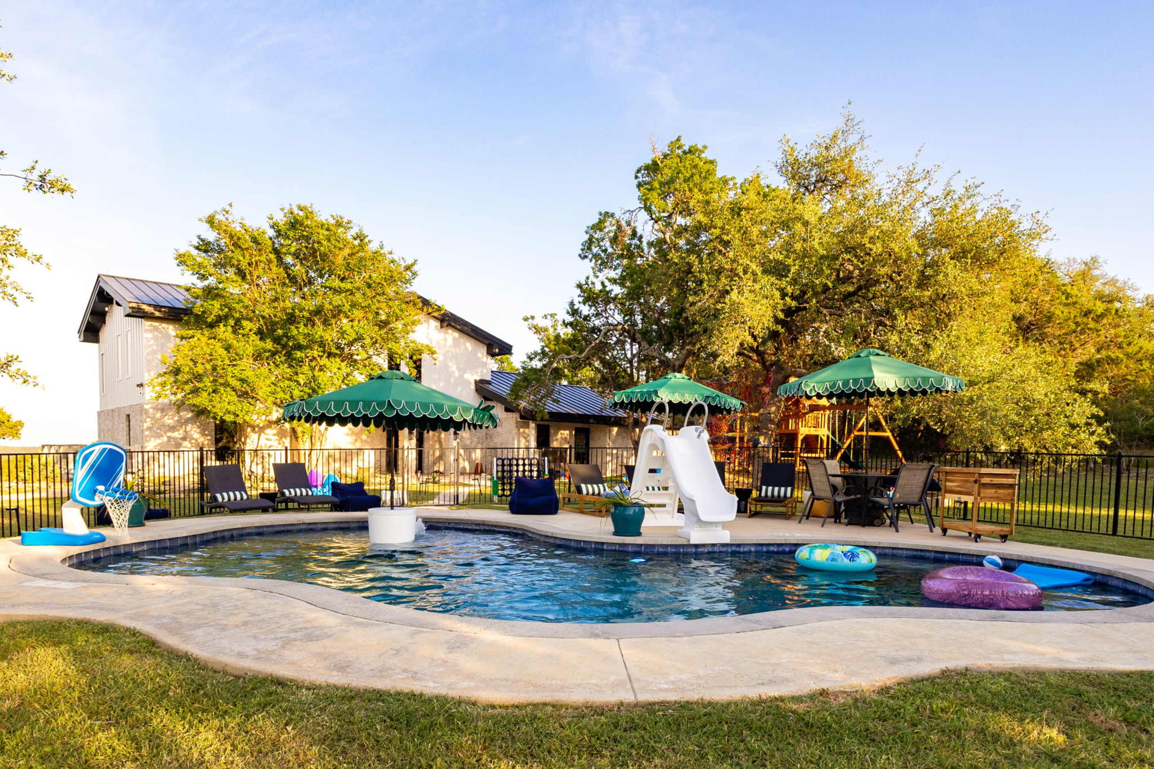 The image shows a backyard pool area with lounge seating, colorful floats, and two green umbrellas, surrounded by a fence and greenery, alongside a house.